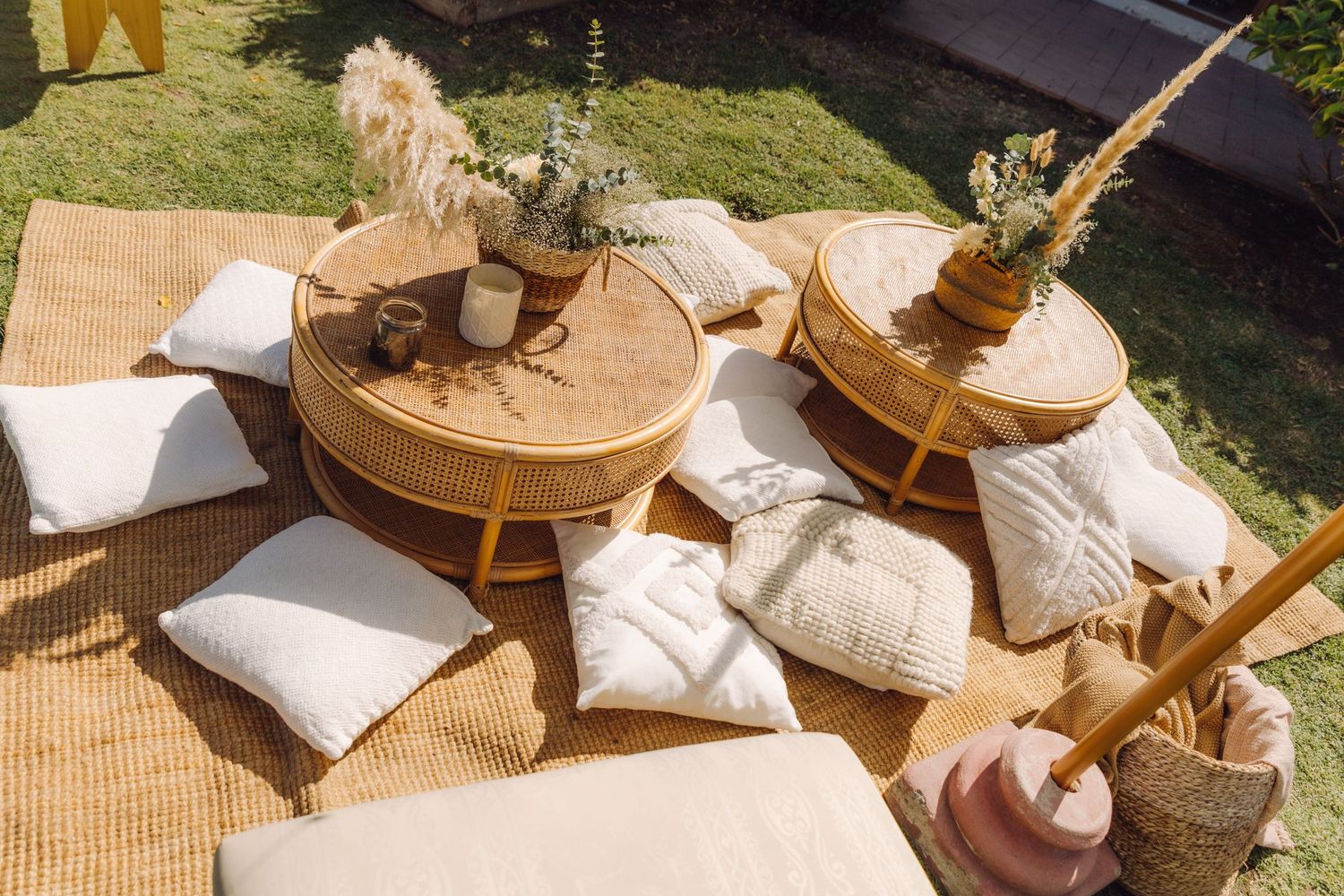 Bohemian outdoor picnic setup with rattan tables, white textured pillows, and dried floral arrangements on a woven mat.