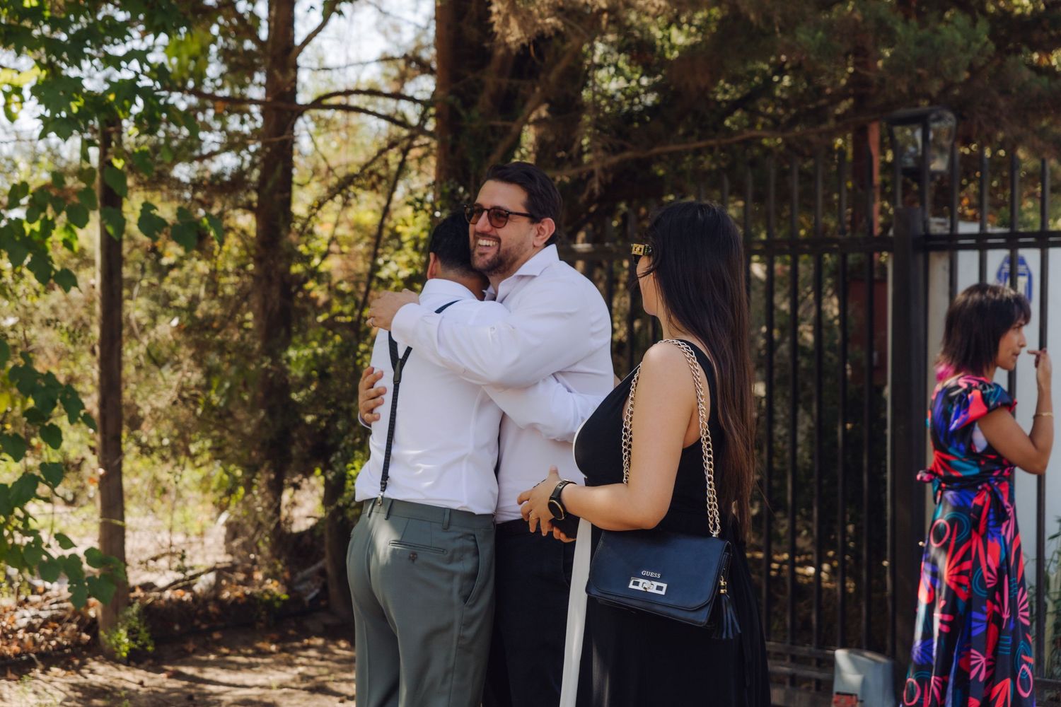Guests greeting and hugging at an outdoor wedding venue near iron gates surrounded by trees.