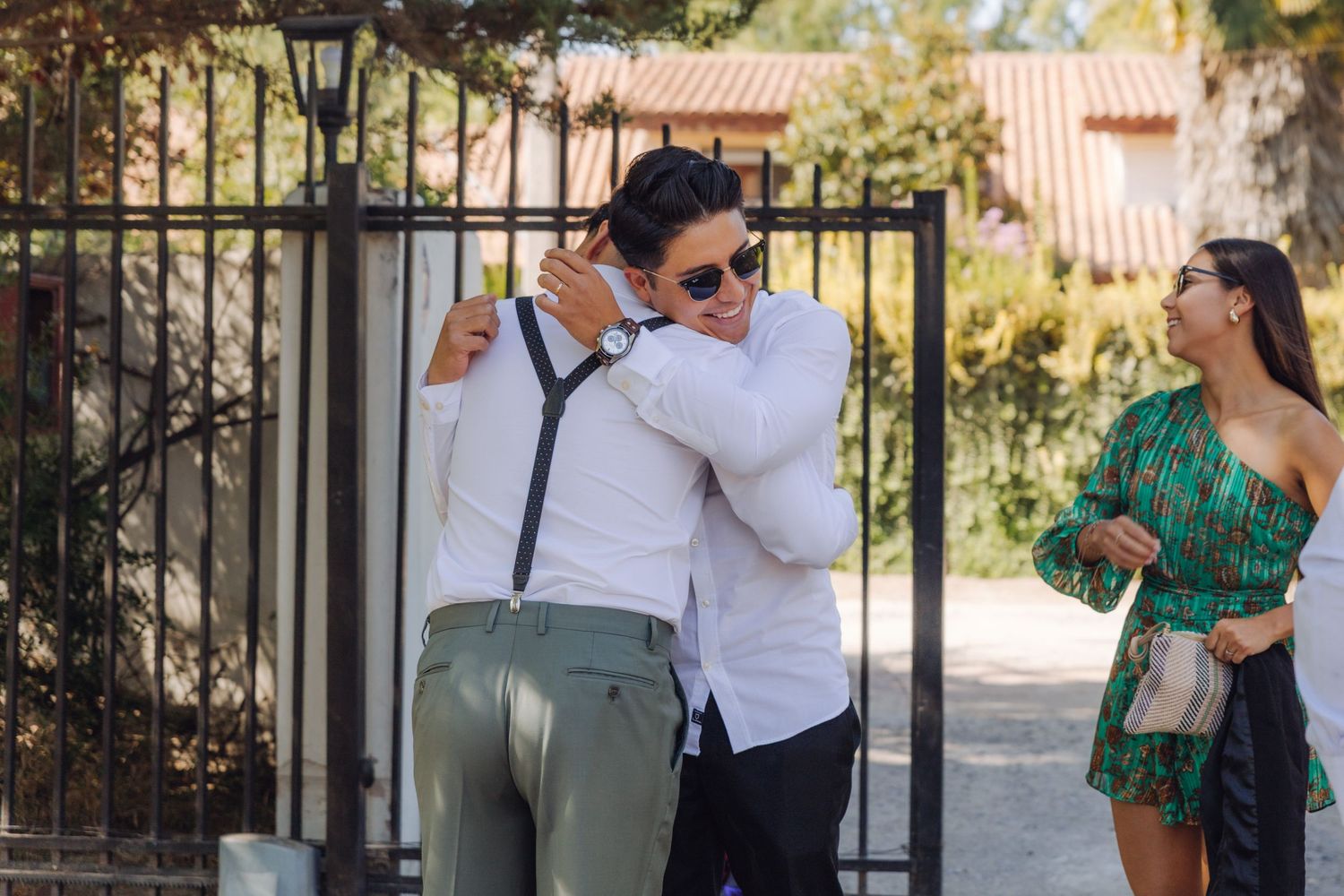 Two men embracing in a warm hug at an outdoor wedding while a woman in green watches nearby.