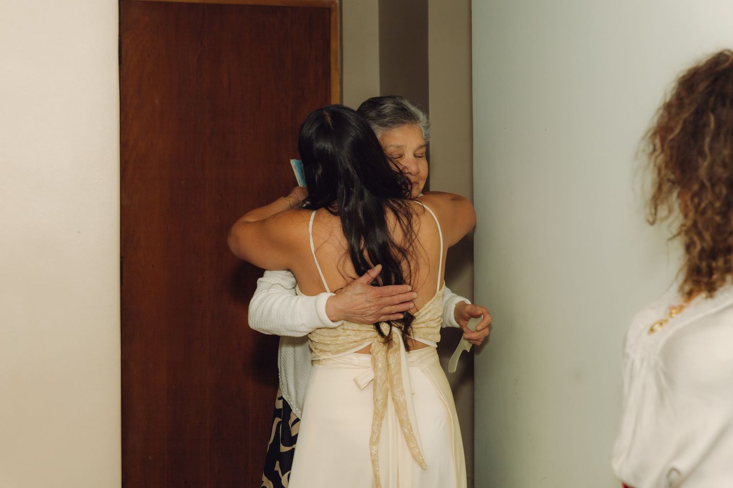 Bride in white wedding dress sharing a heartfelt embrace with an older woman in a doorway.