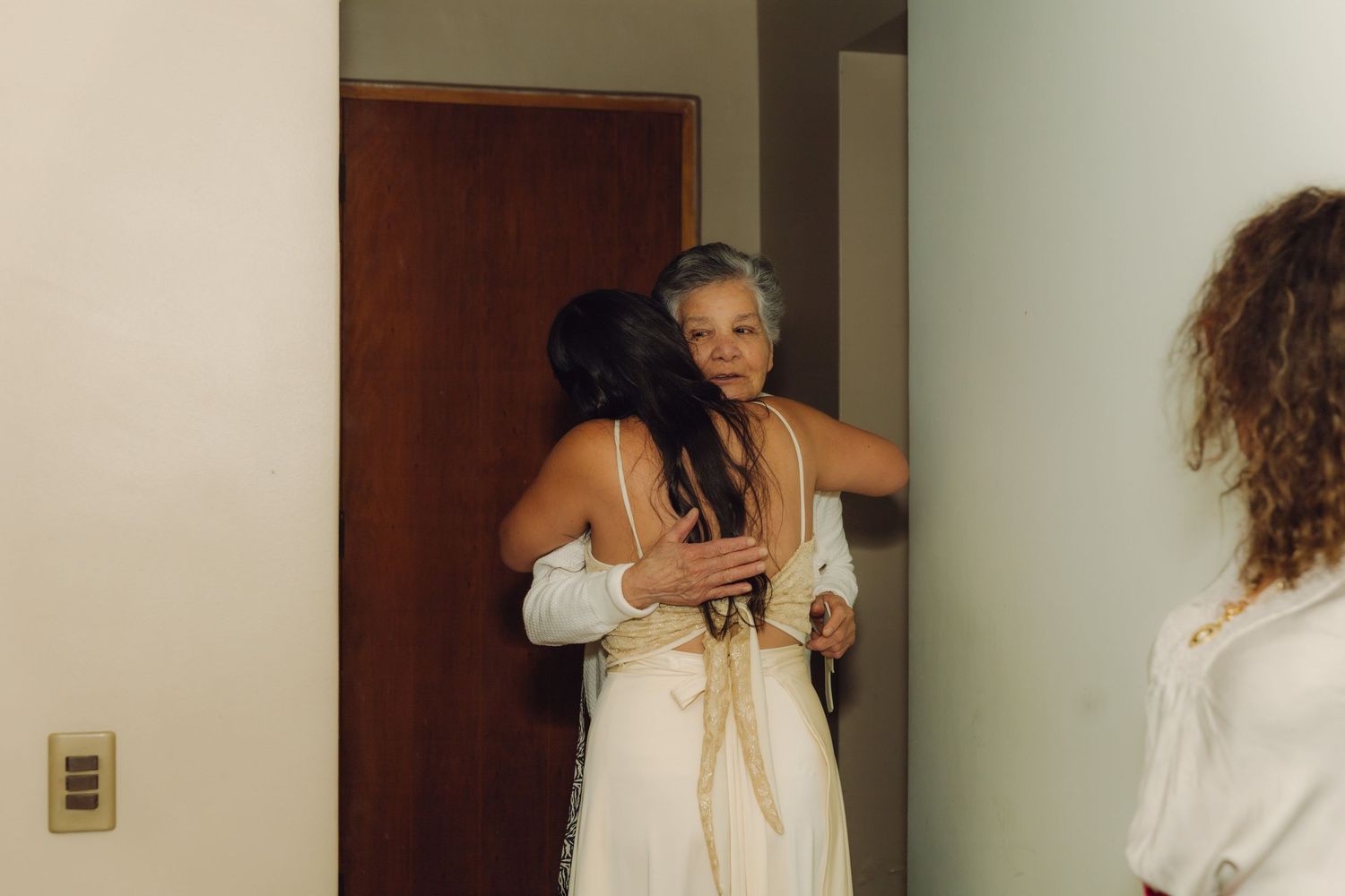 Bride in elegant white gown hugging an older woman affectionately in a bright indoor hallway.