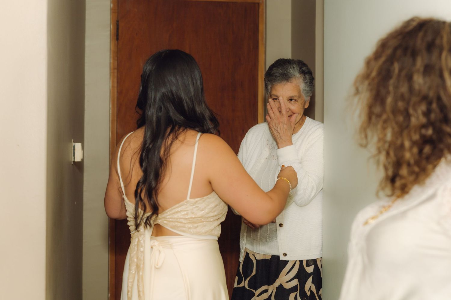 Older woman emotionally covering her mouth while admiring the bride in a white wedding dress indoors.