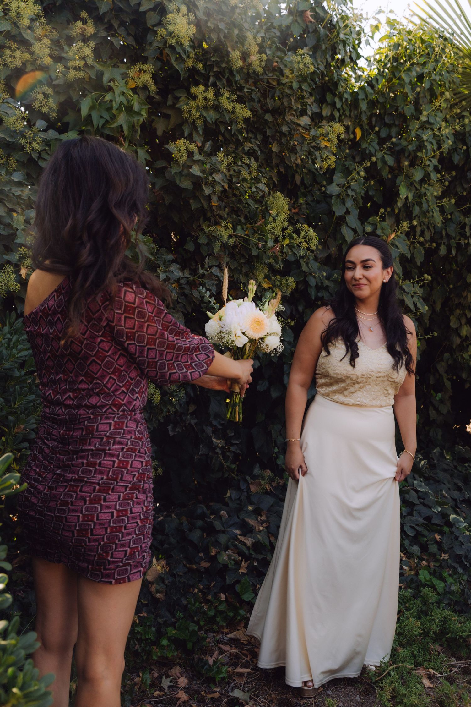 Bridesmaid in red dress handing a white rose bouquet to the bride in a cream gown outdoors.