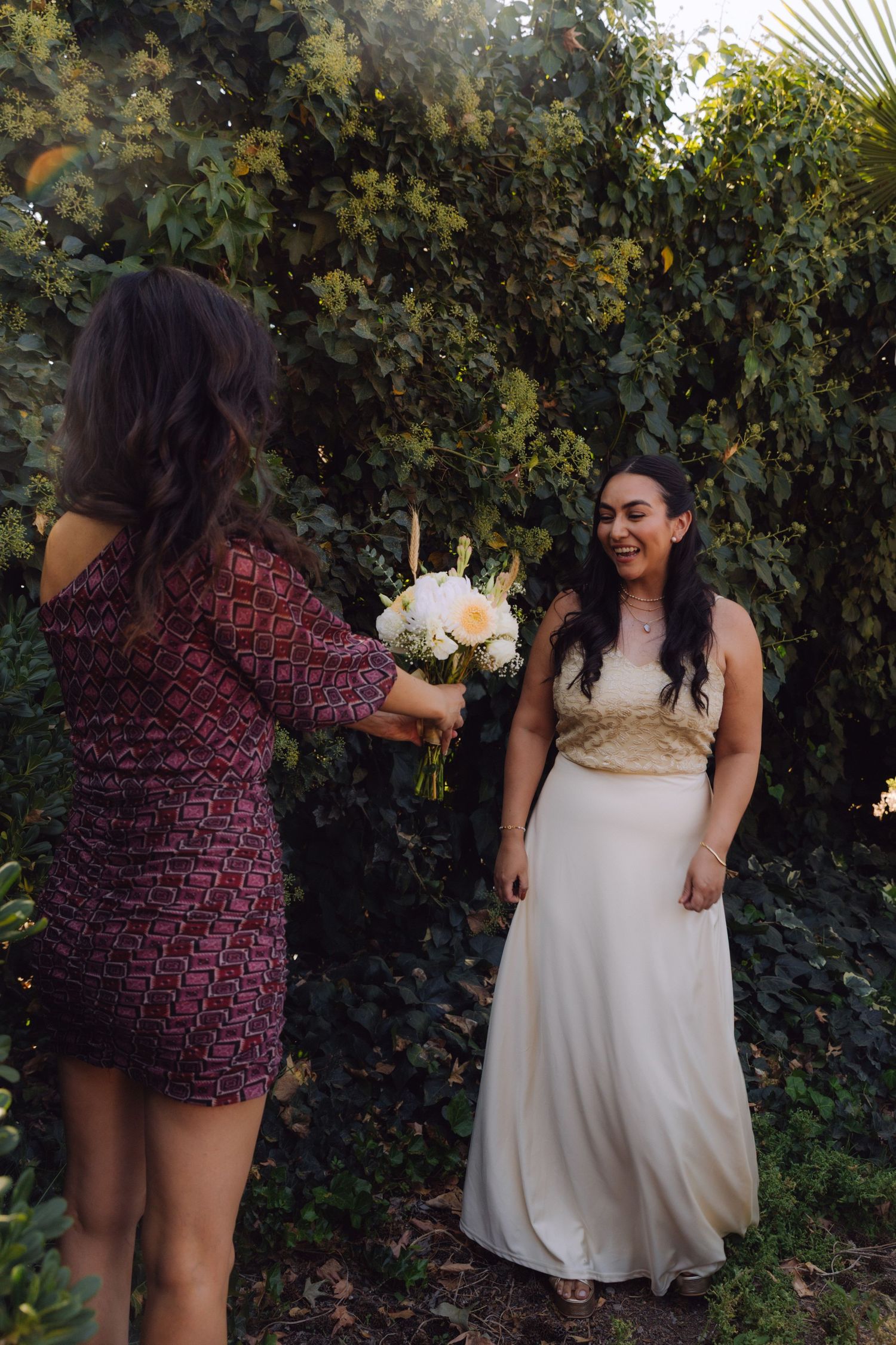 Bride laughing joyfully as friend presents her with a white floral bouquet outdoors.
