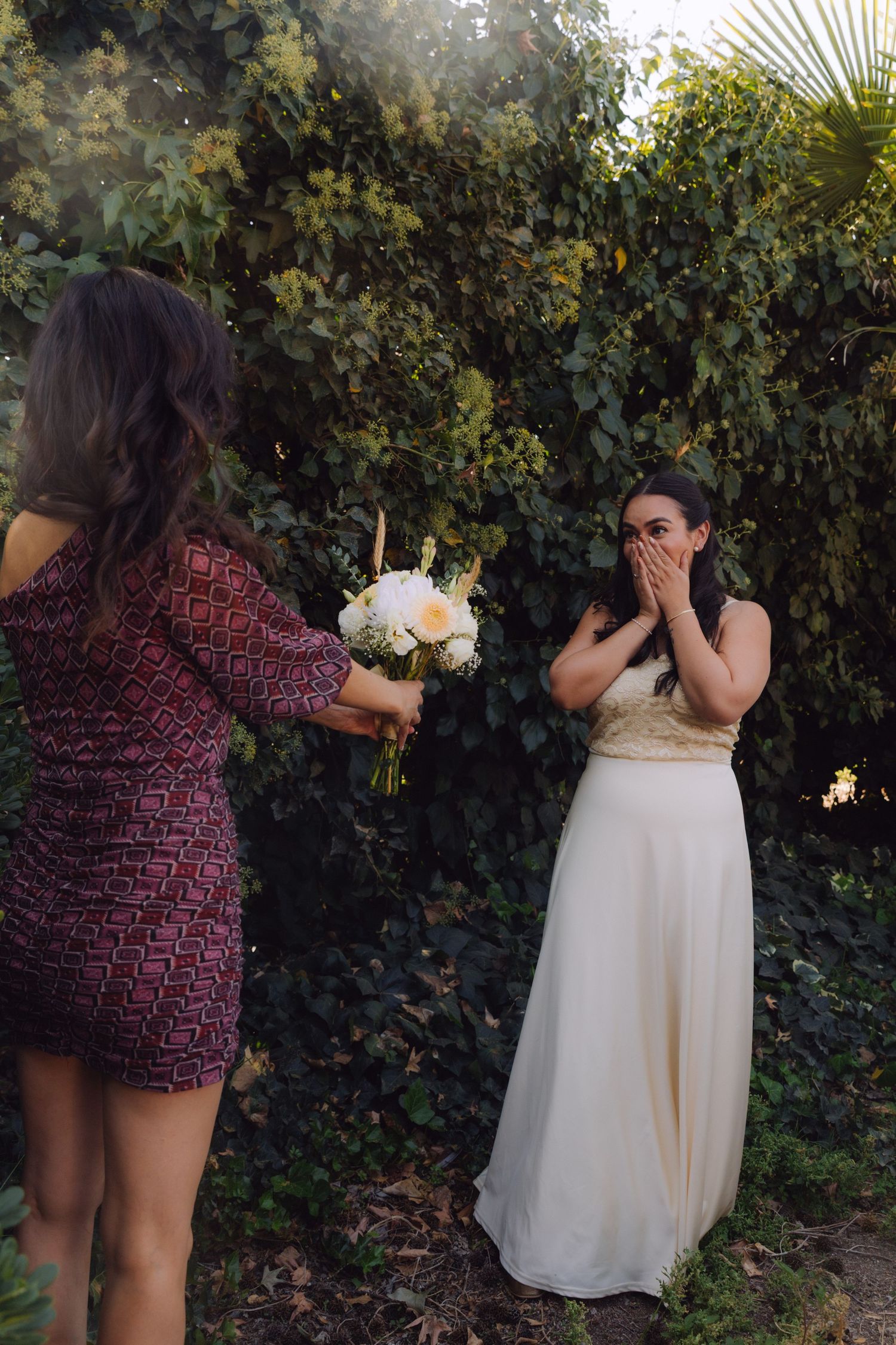 Emotional bride covering her mouth in surprise as friend offers her a wedding bouquet outdoors.