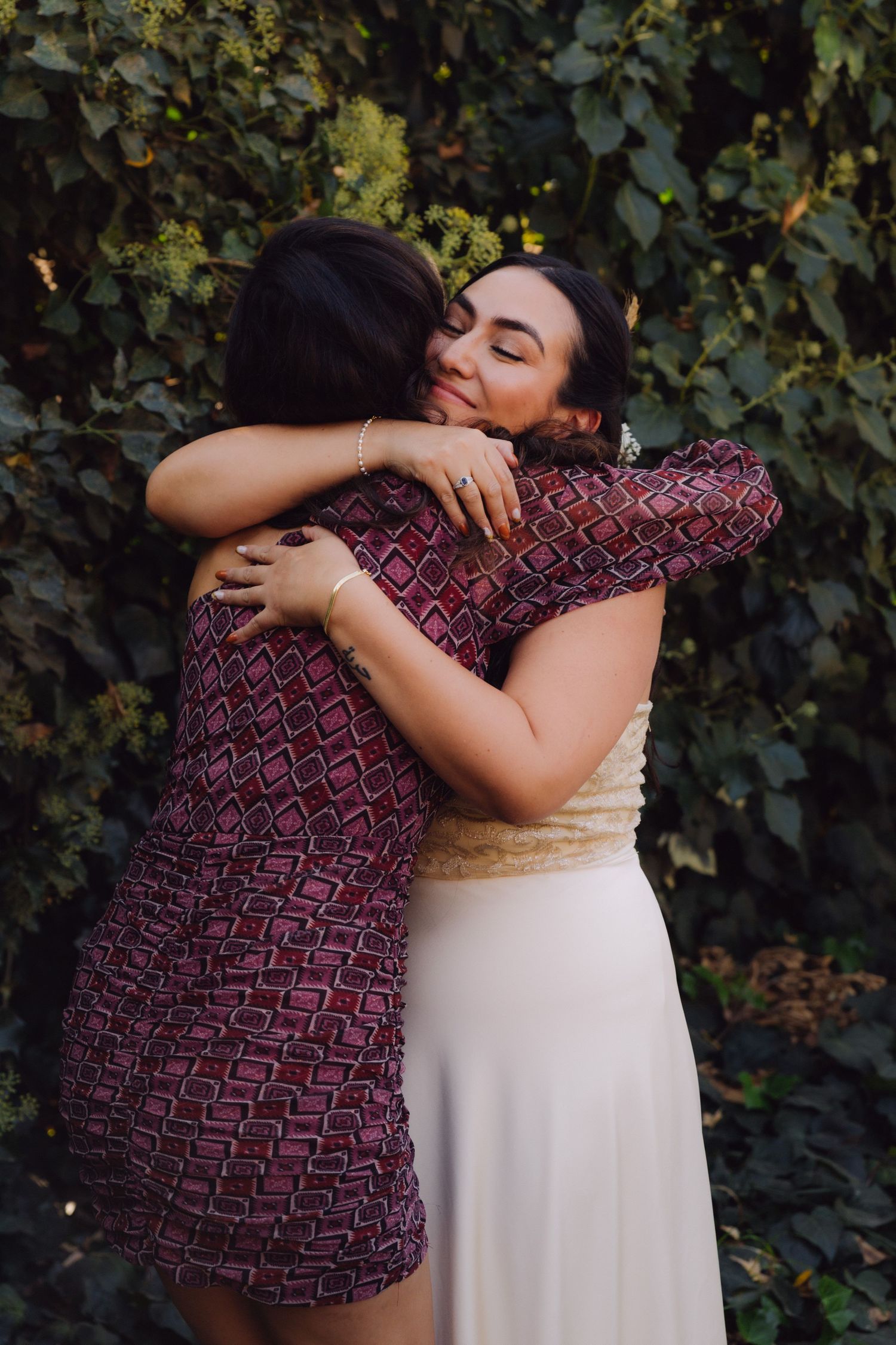 Bride and friend sharing a warm emotional hug in front of lush green foliage.