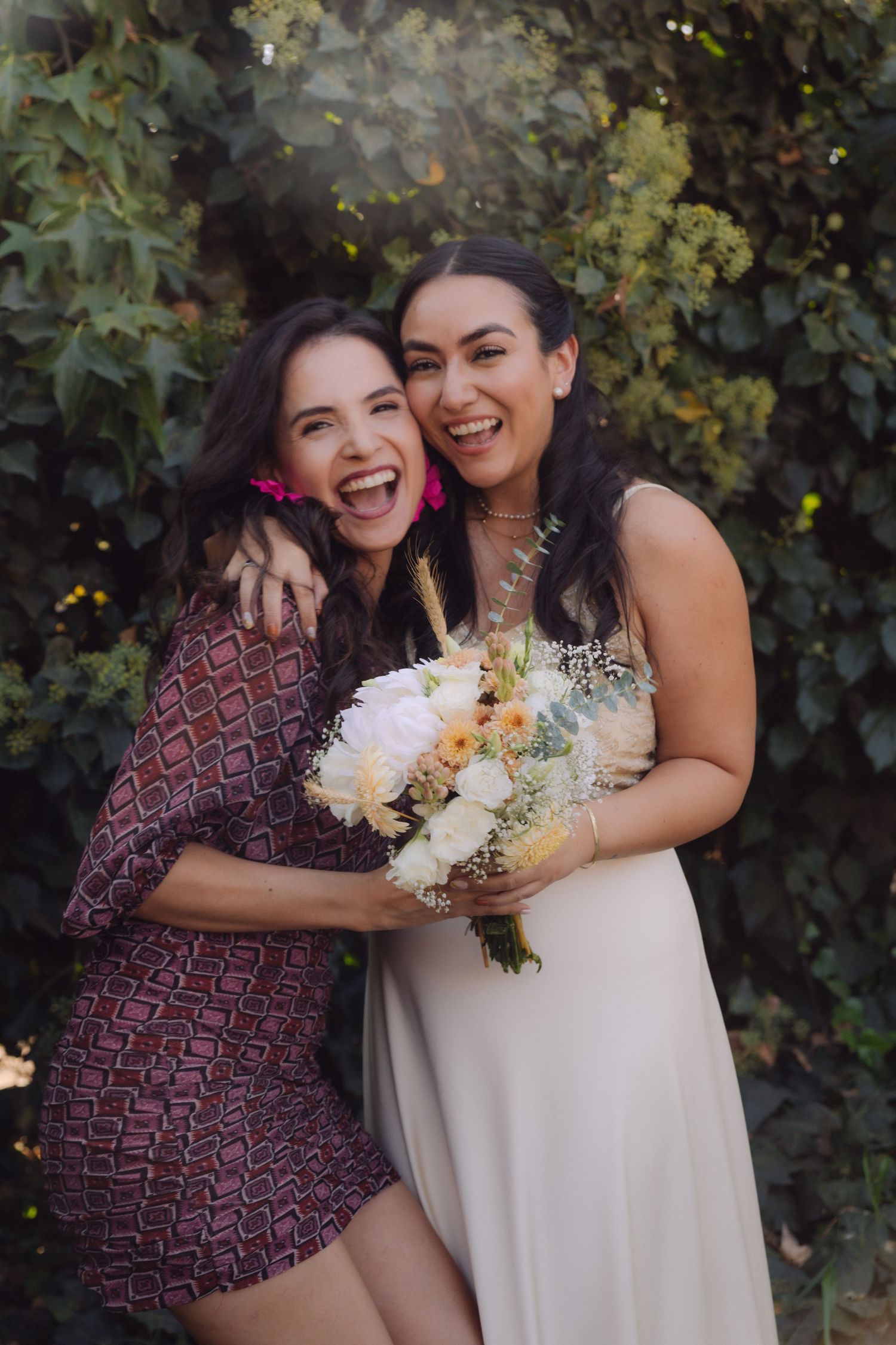 Laughing bride and friend posing together holding a white and peach floral bouquet outdoors.