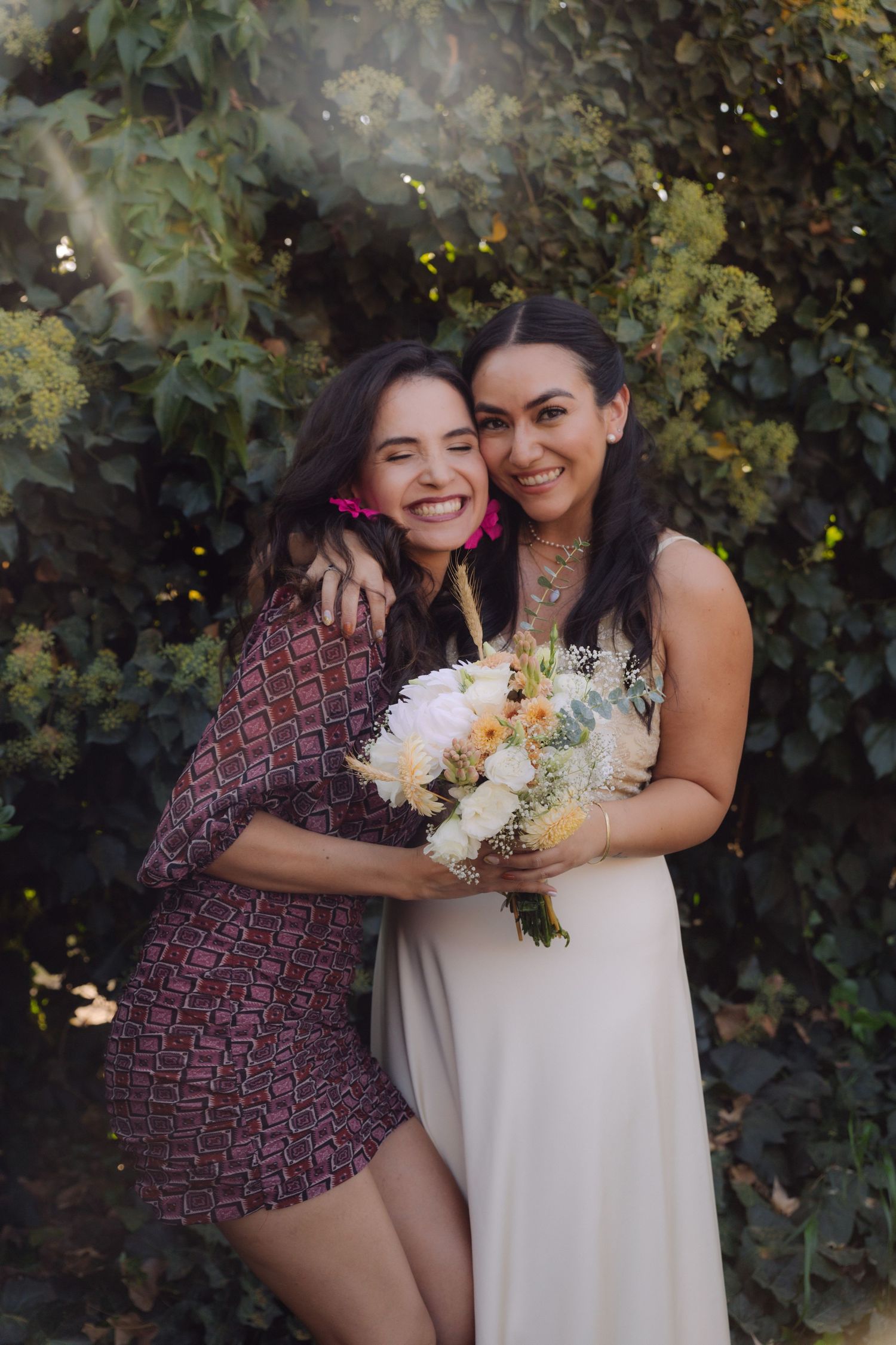 Happy bride holding bouquet embracing smiling friend with pink earrings in garden setting.