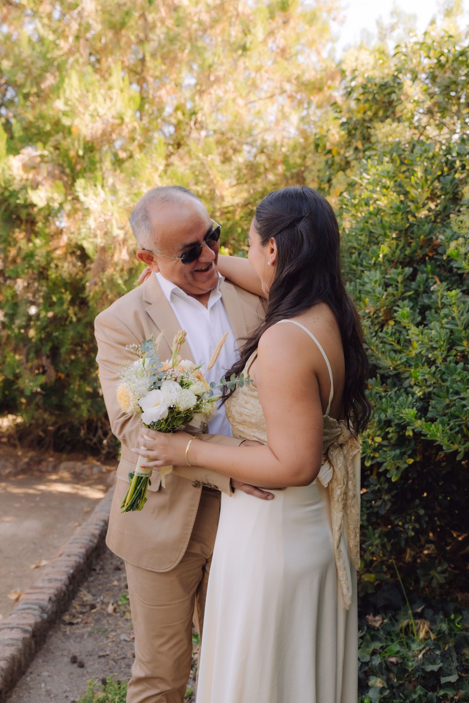 Bride and older man in tan suit sharing a tender moment holding a white bouquet outdoors.