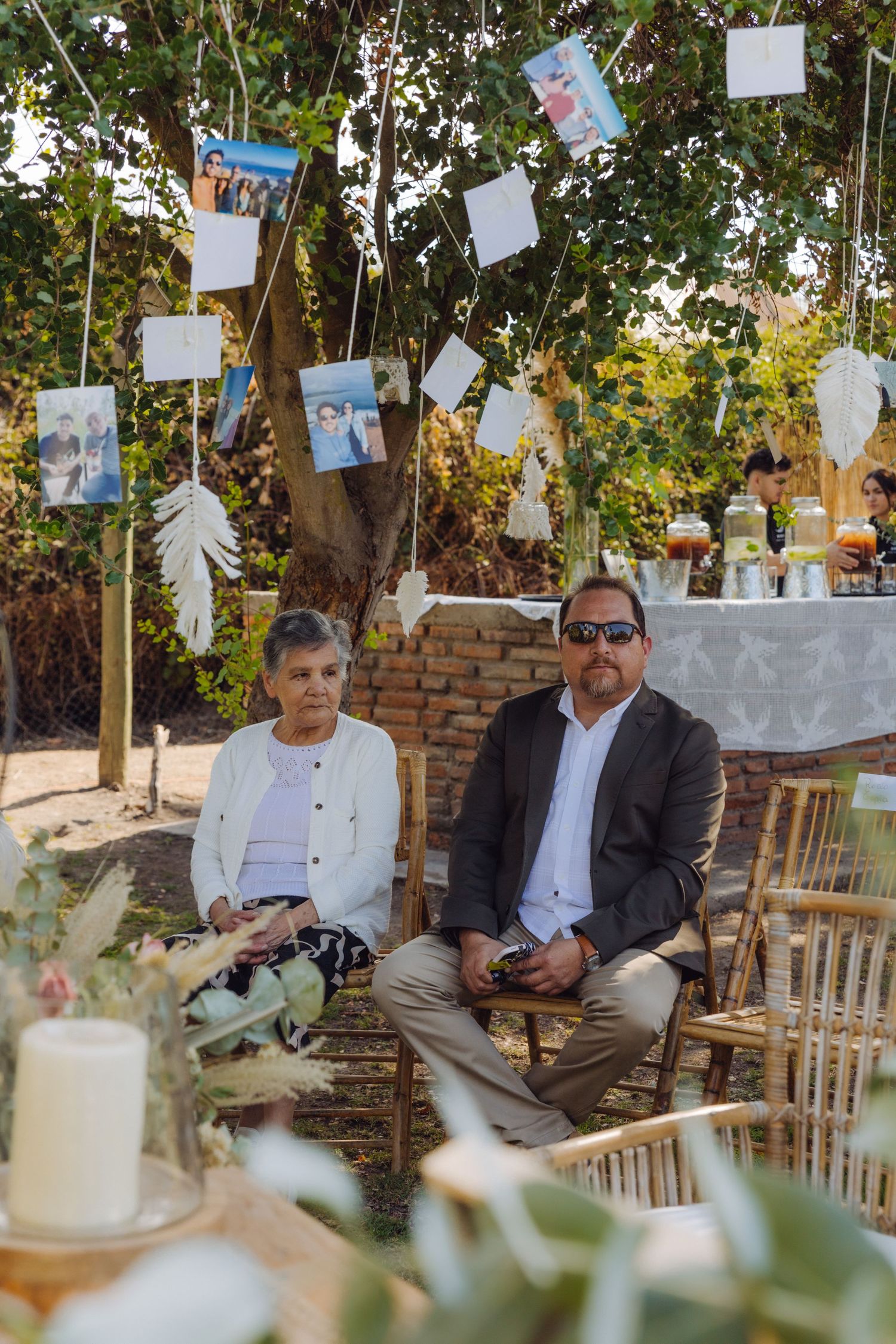 Elderly couple seated at an outdoor wedding reception beneath hanging photo decorations.
