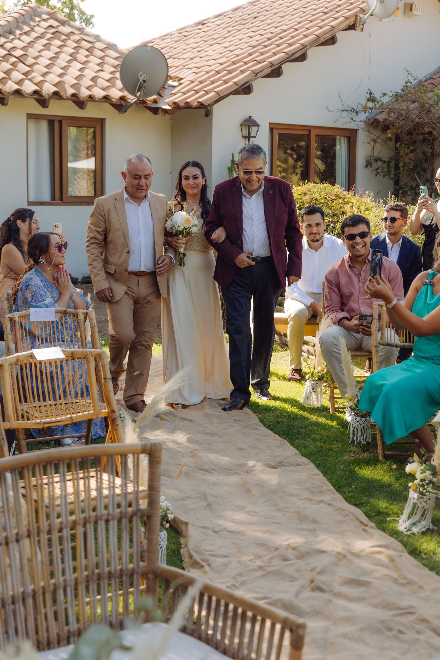 Bride walking down sandy outdoor aisle escorted by two men at a garden wedding ceremony.