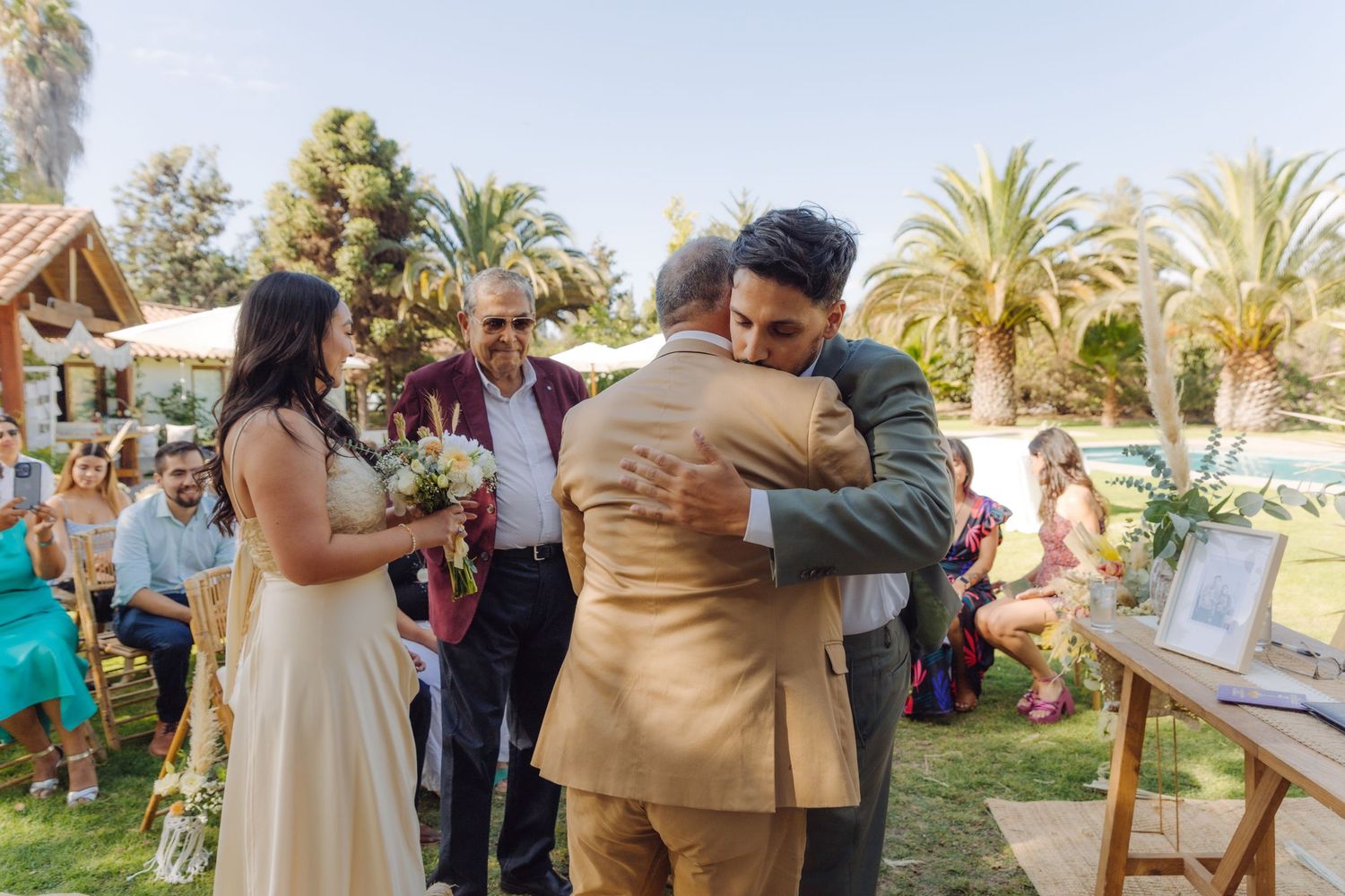 Groom embracing father figure at outdoor wedding altar as bride stands nearby holding bouquet.