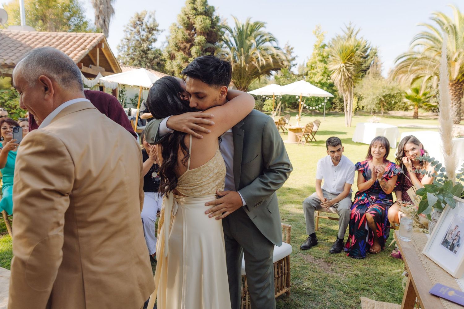 Bride and groom sharing first kiss at outdoor wedding ceremony with guests watching nearby.