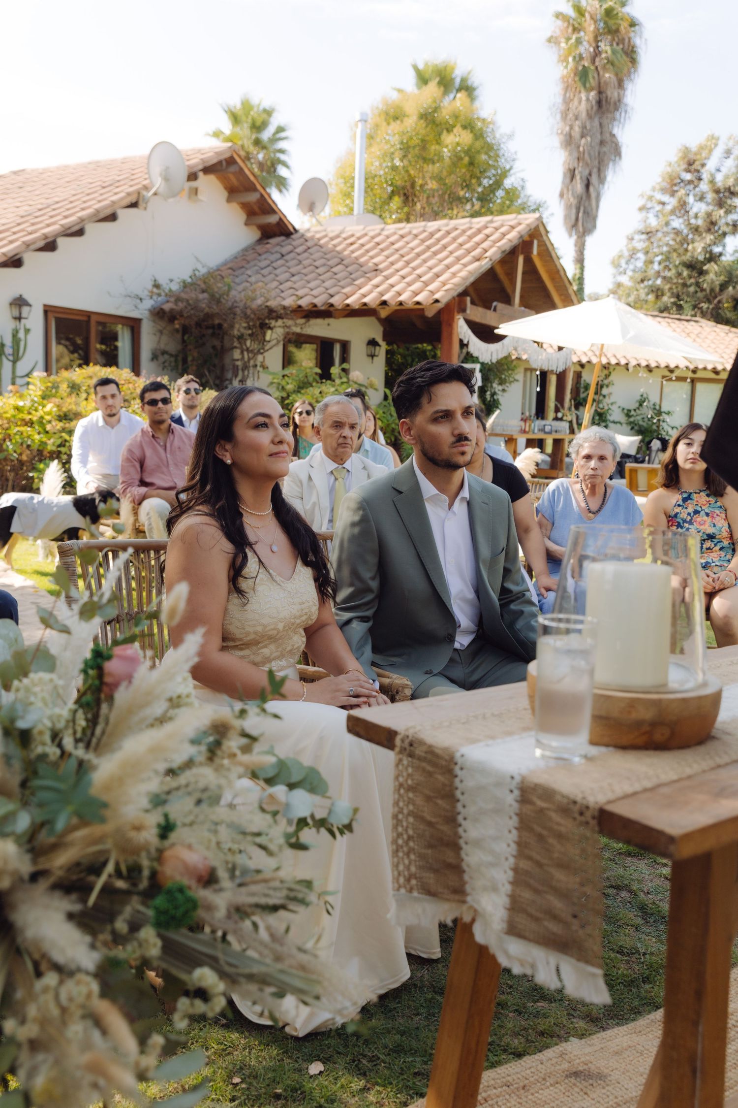 Bride and groom sit together during outdoor wedding ceremony surrounded by floral decorations.