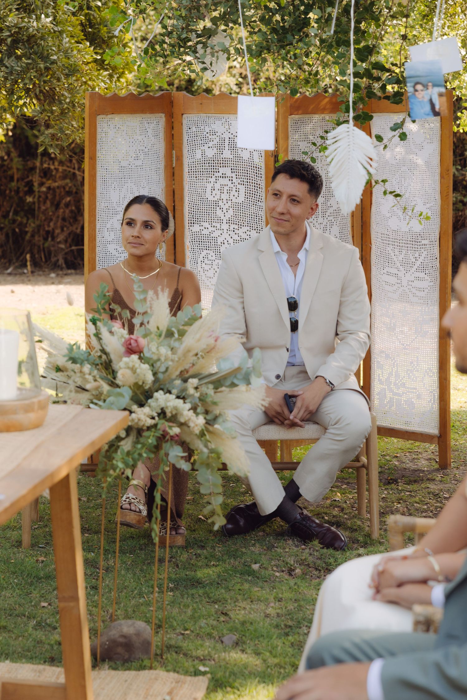 Couple sits together at outdoor boho wedding ceremony with lace screen backdrop and floral arrangements.