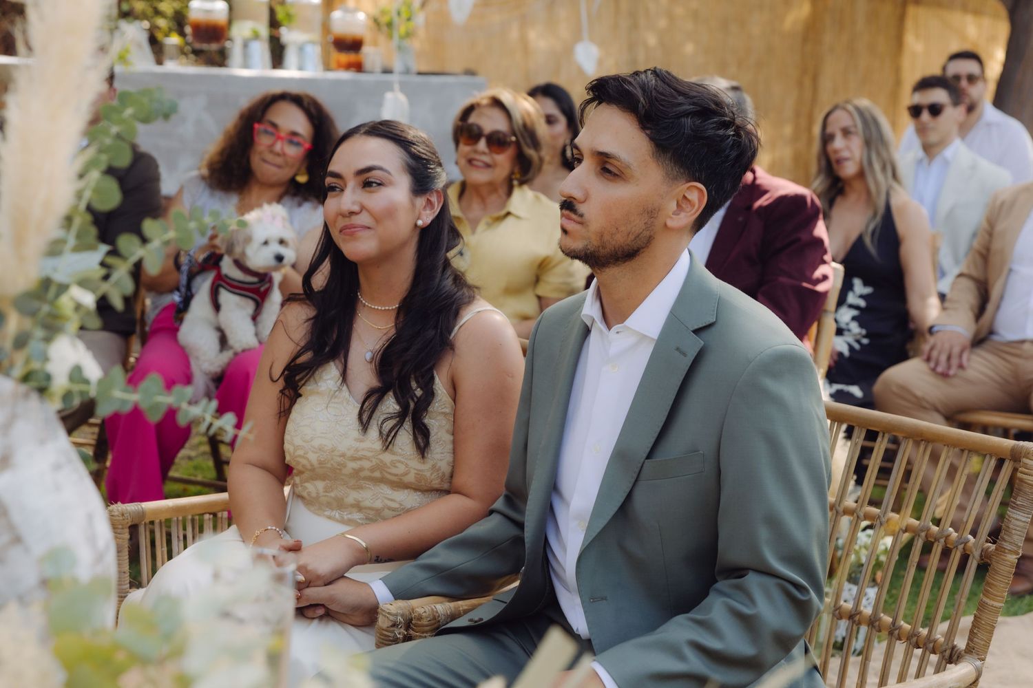 Bride and groom attentively seated during outdoor wedding ceremony with guests gathered behind them.