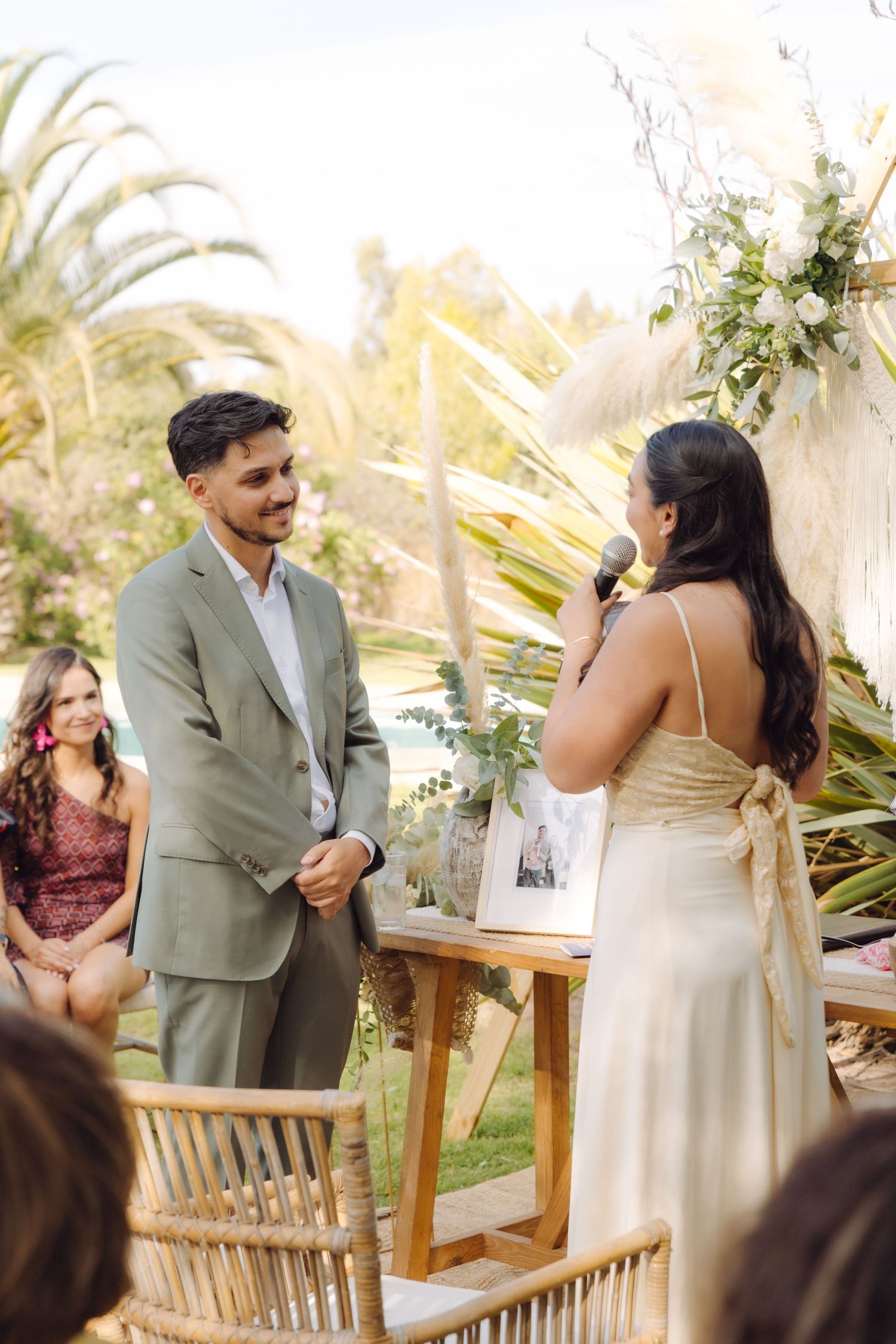 Bride reads vows to groom at outdoor boho wedding ceremony surrounded by tropical greenery and floral arrangements.