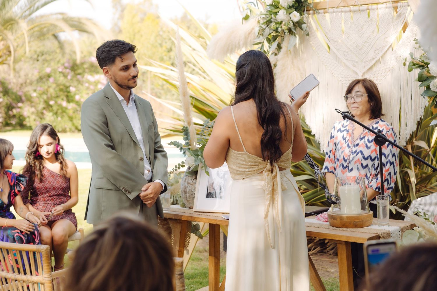 Couple exchanges vows at elegant outdoor wedding ceremony with macrame backdrop and lush tropical palm decorations.