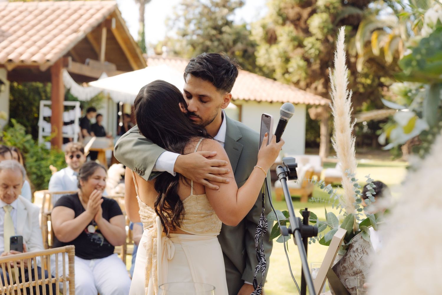 Newlyweds share emotional first embrace after wedding ceremony in beautiful outdoor garden venue with guests applauding.