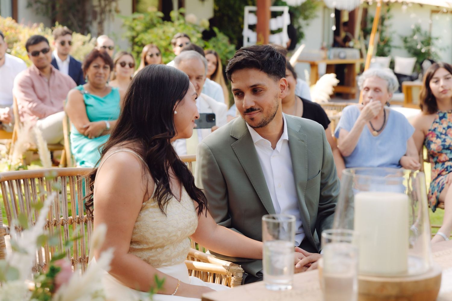 Bride and groom sit together at wedding reception table sharing an intimate moment with guests seated behind them.