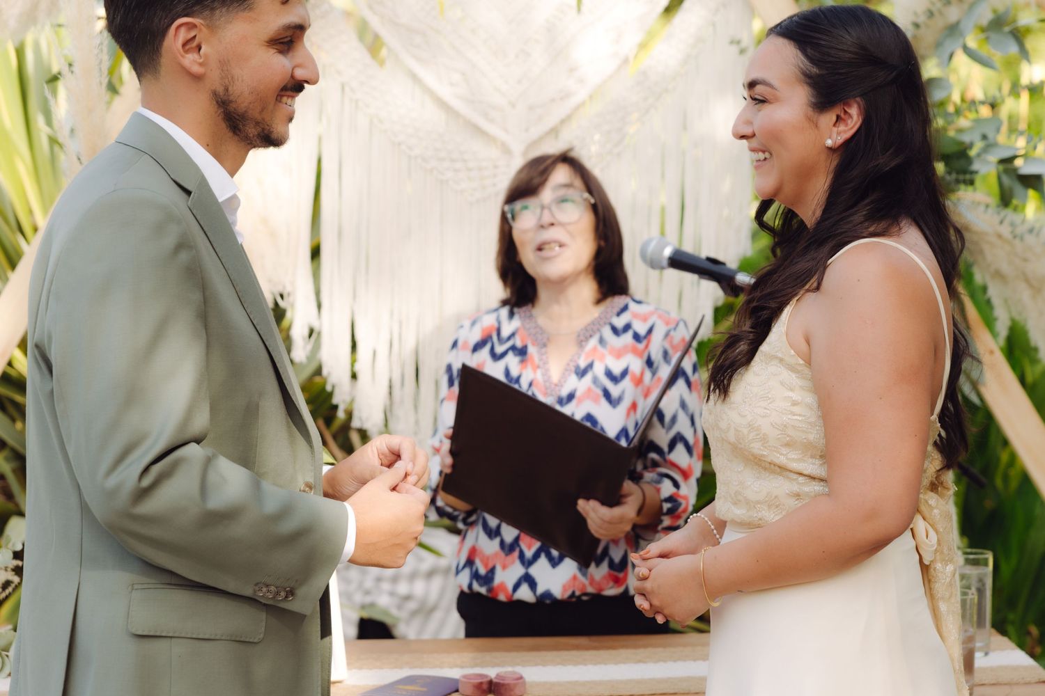 Bride and groom exchange rings during outdoor wedding ceremony officiated by woman in colorful blazer with macrame backdrop.