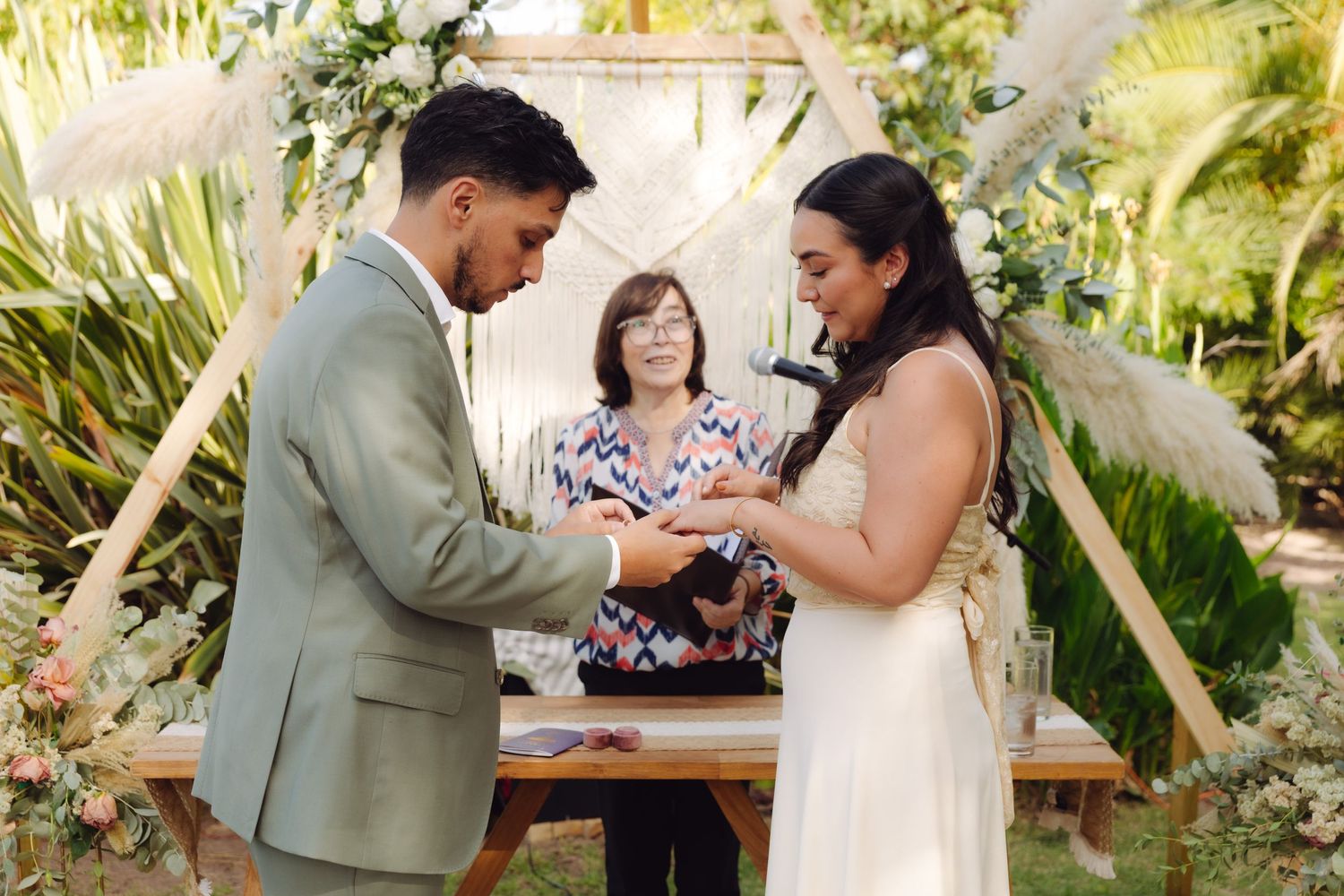 Couple exchanges wedding rings during outdoor boho ceremony with officiant and geometric wooden arch with floral decor.