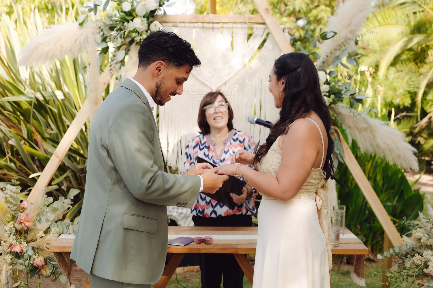 Groom places wedding ring on brides finger during outdoor ceremony with officiant and tropical greenery backdrop.