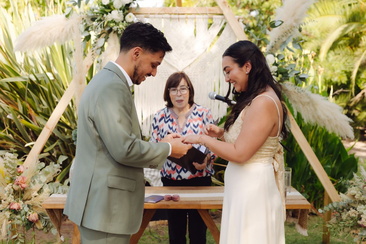 Bride and groom exchange wedding rings at outdoor ceremony with smiling officiant and elegant floral arch behind them.