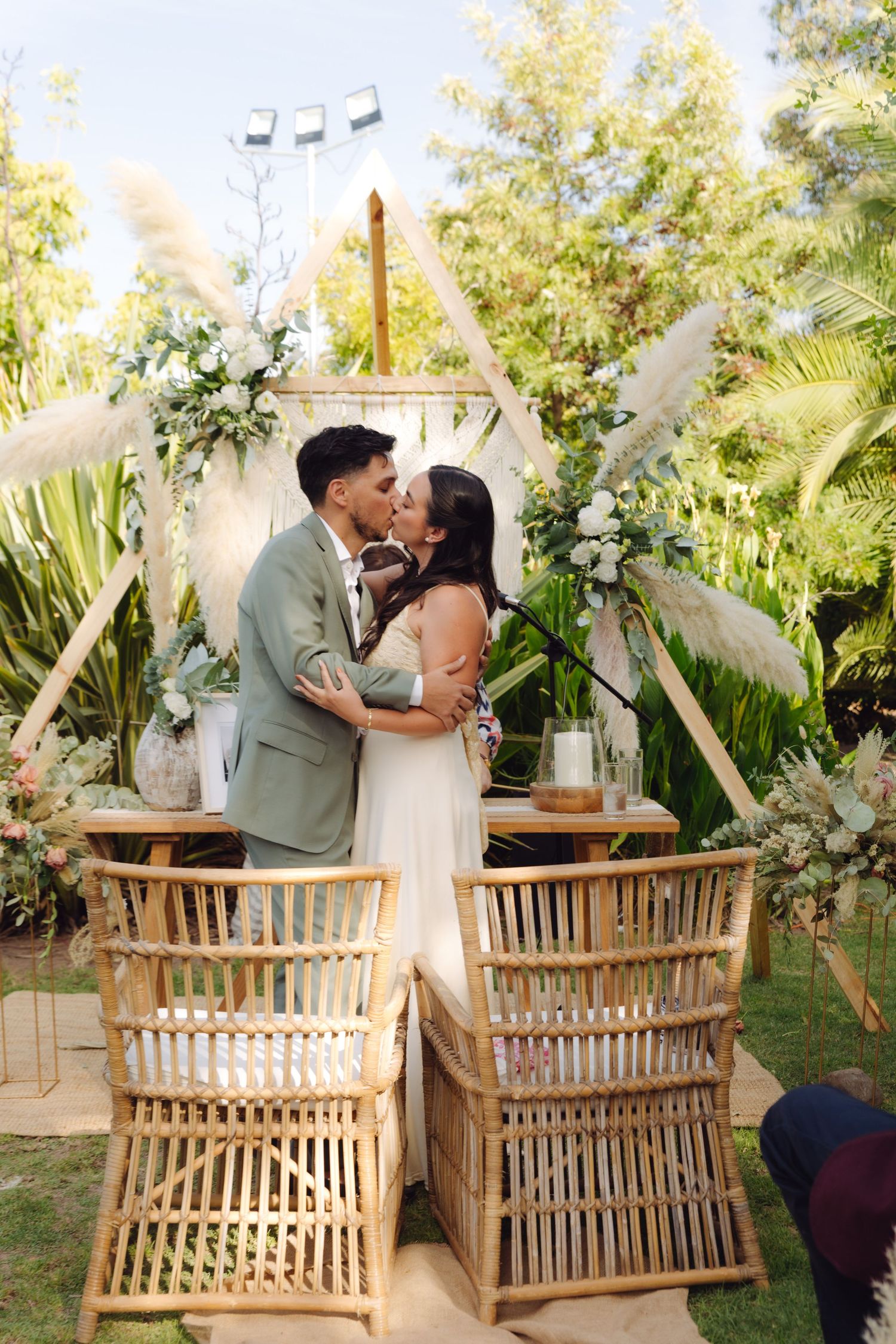 Newlyweds share first kiss at outdoor boho wedding altar with geometric arch, pampas grass, and white floral arrangements.
