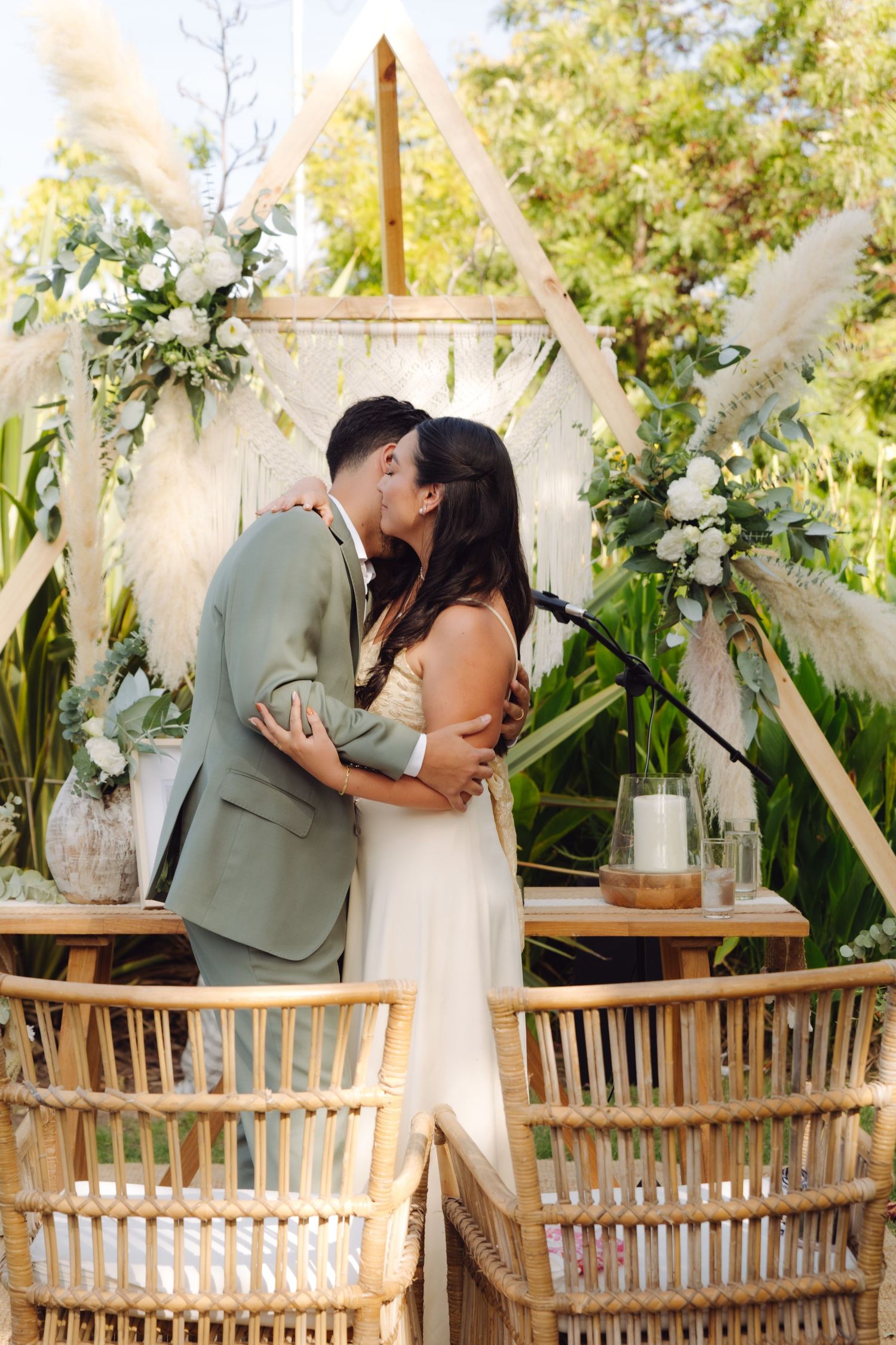 Bride and groom share their first kiss at an outdoor boho wedding ceremony with a geometric wooden arch and pampas grass.
