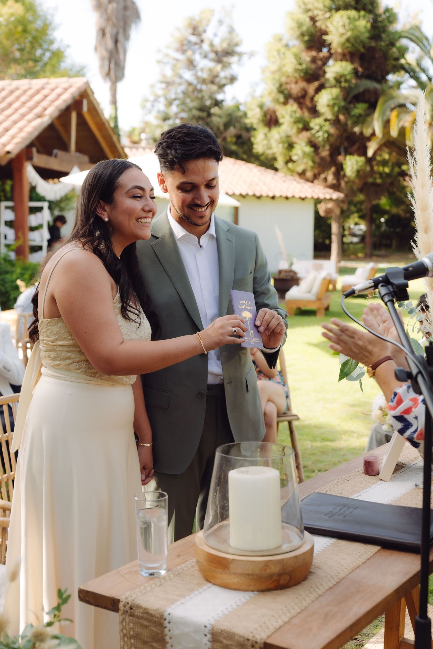 Smiling bride and groom participate in a unity ceremony at an outdoor wedding with a candle and decorative table.
