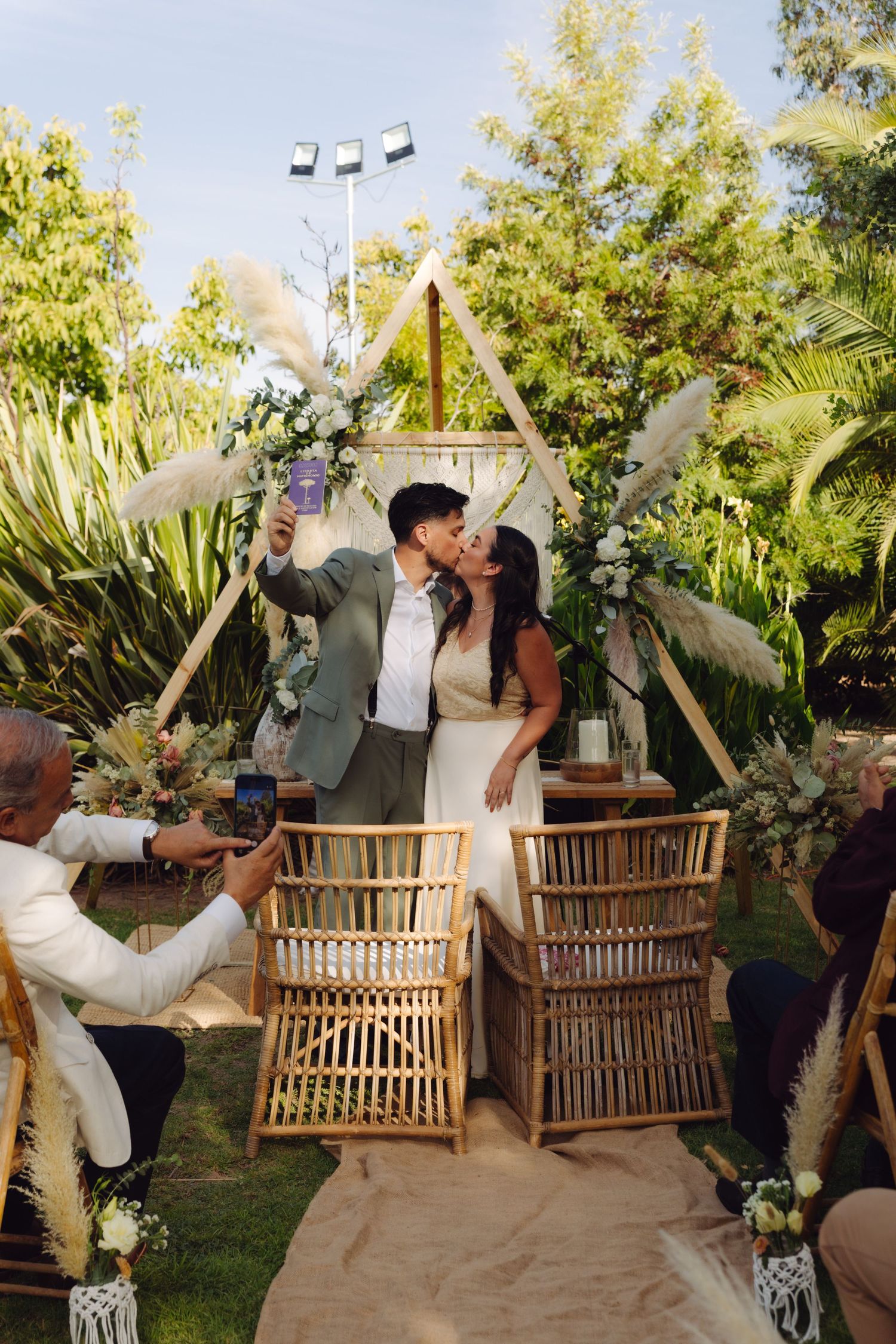 Newlyweds kiss and take a selfie at their outdoor boho wedding ceremony with a triangular wooden arch backdrop.