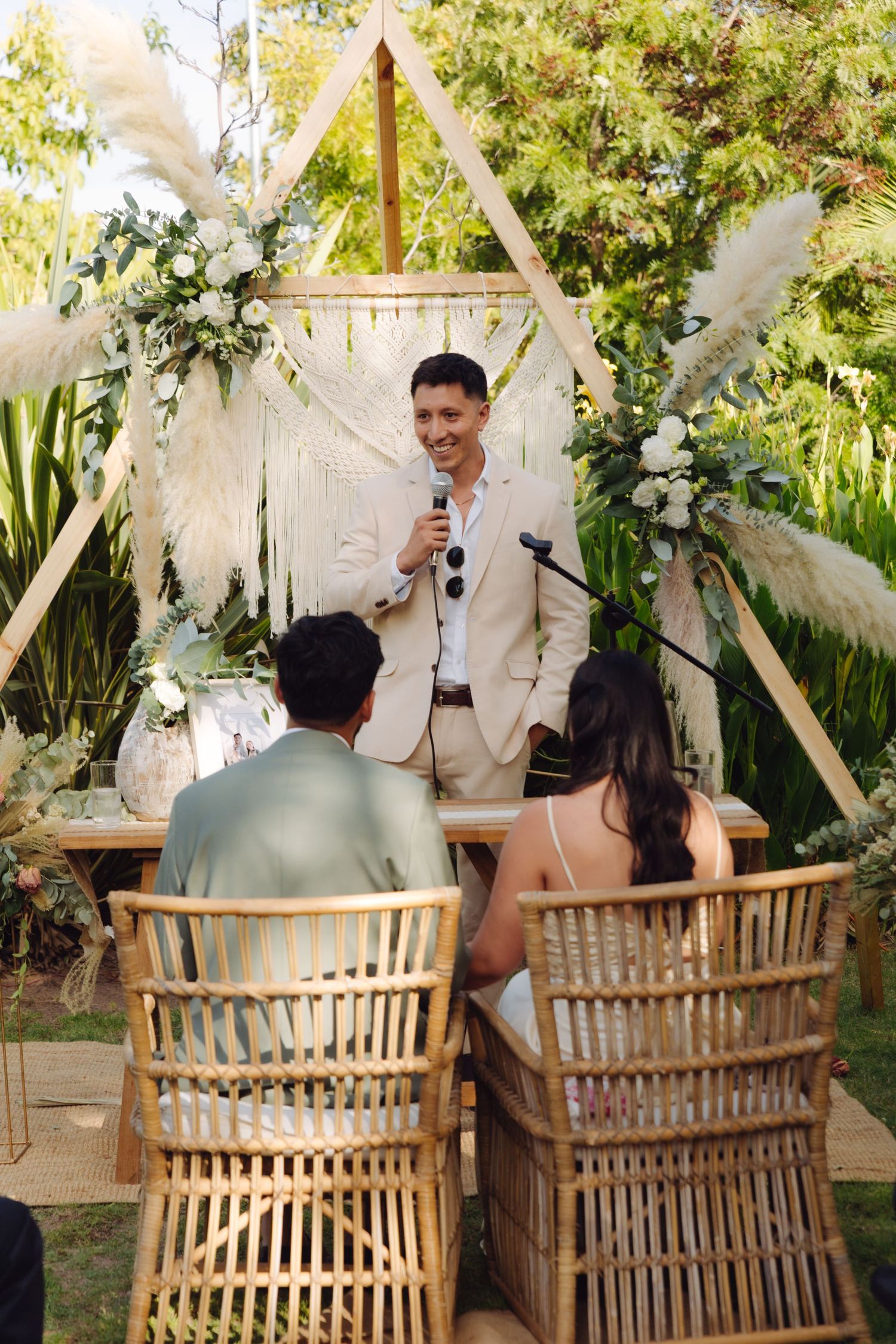 Man in beige suit speaks at microphone during an outdoor boho wedding ceremony.
