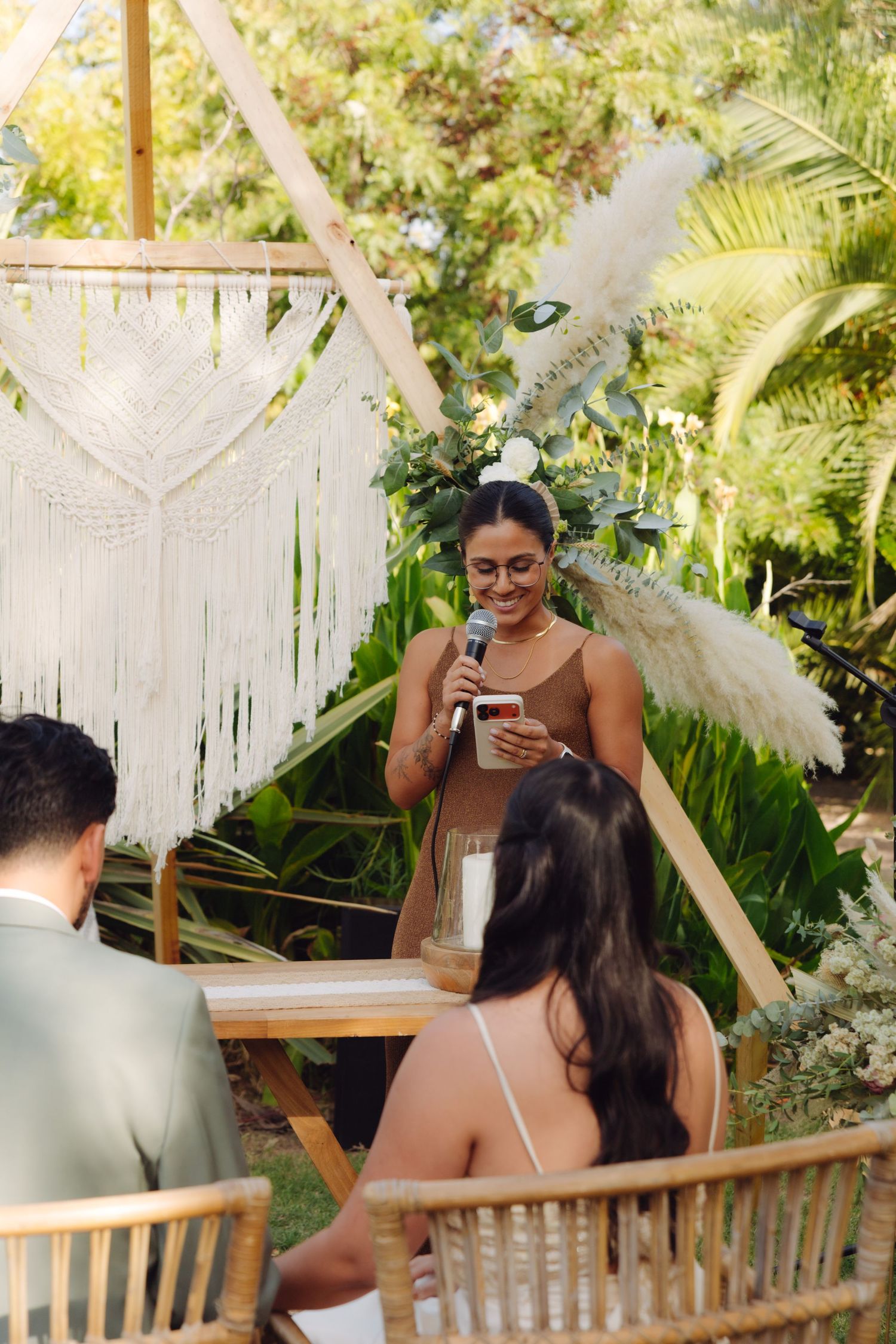 Woman in brown dress reads from phone at microphone during outdoor wedding ceremony.
