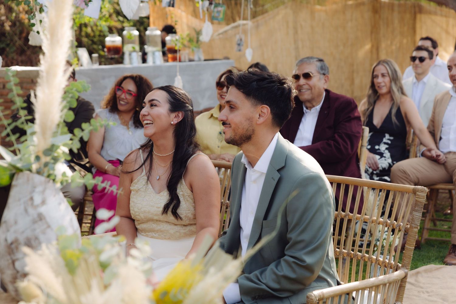 Smiling bride and groom sit together during their outdoor boho wedding ceremony.