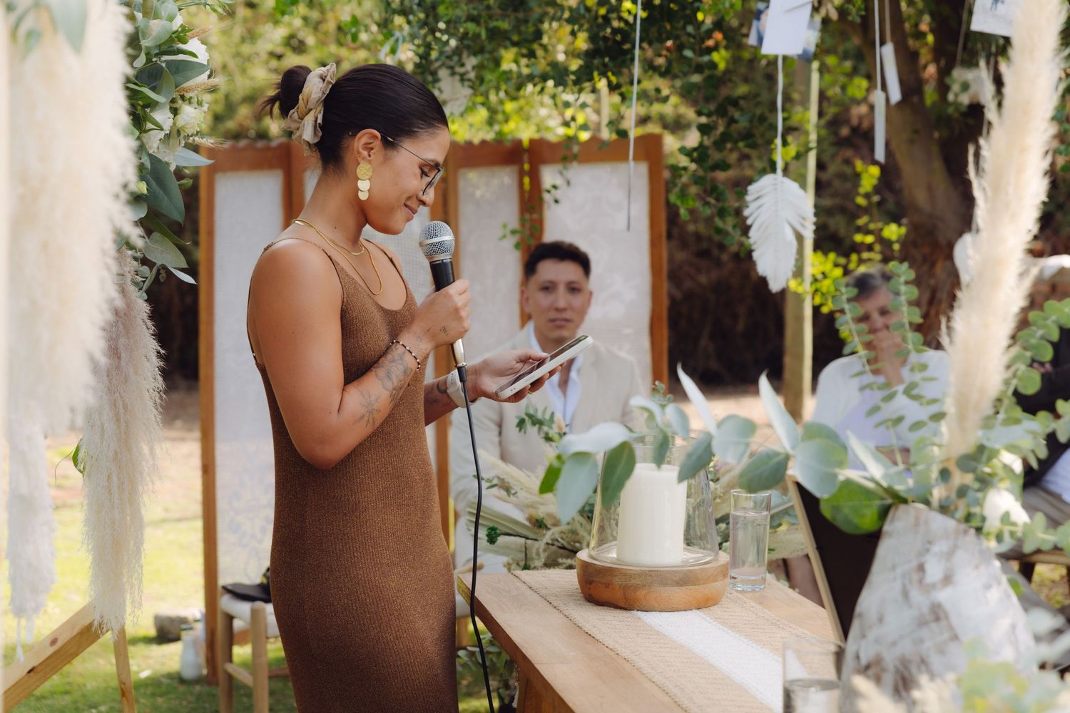 Woman in brown dress speaks into microphone at a decorated outdoor wedding ceremony.