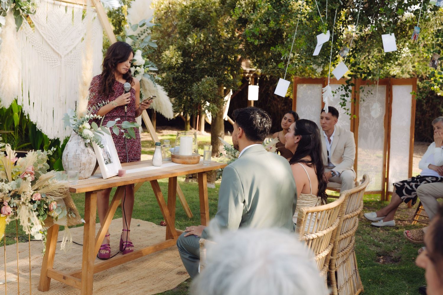 Woman giving a speech at an outdoor boho wedding ceremony with floral decorations and seated guests.