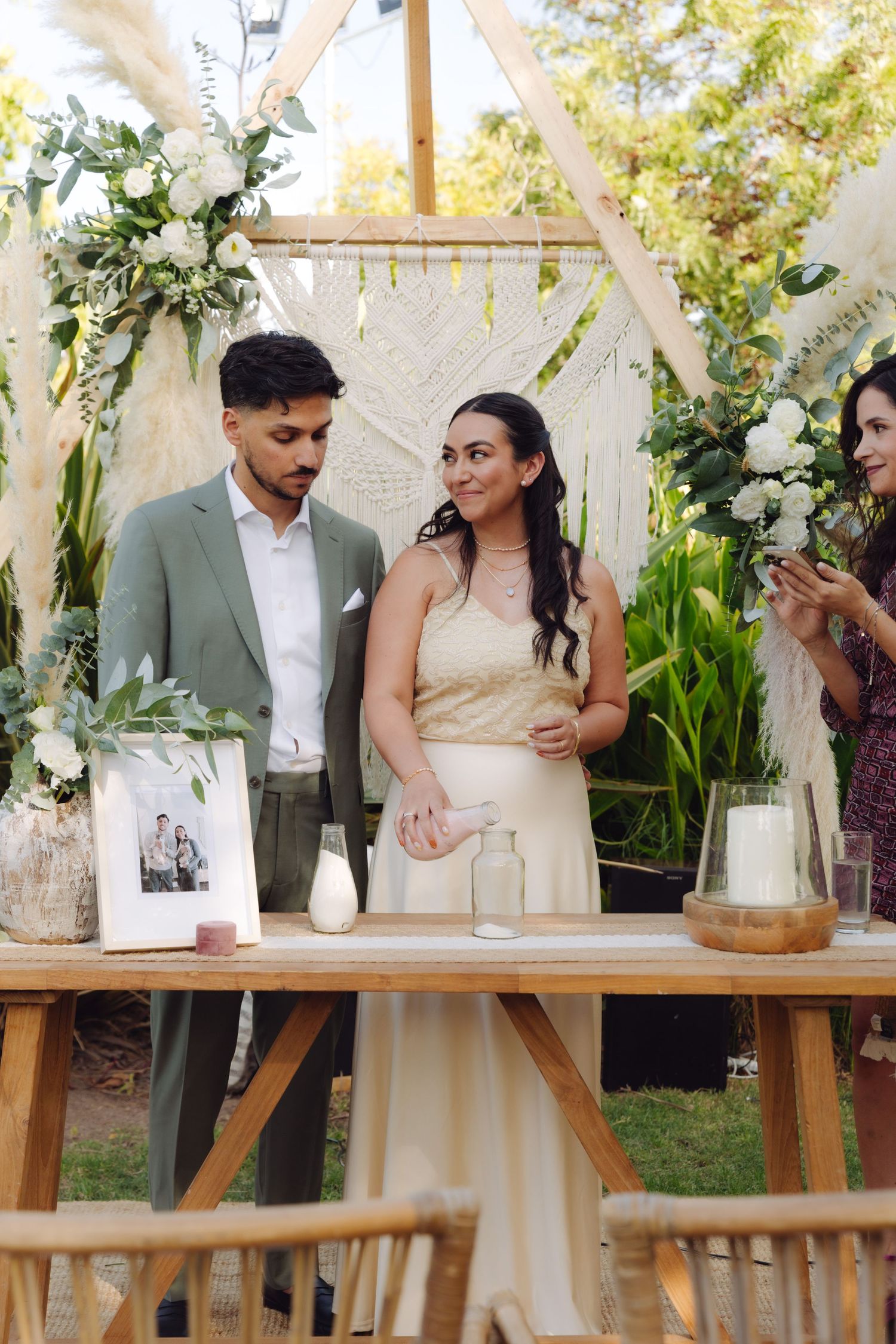 Bride laughing joyfully beside groom at decorated wooden altar in outdoor boho wedding.