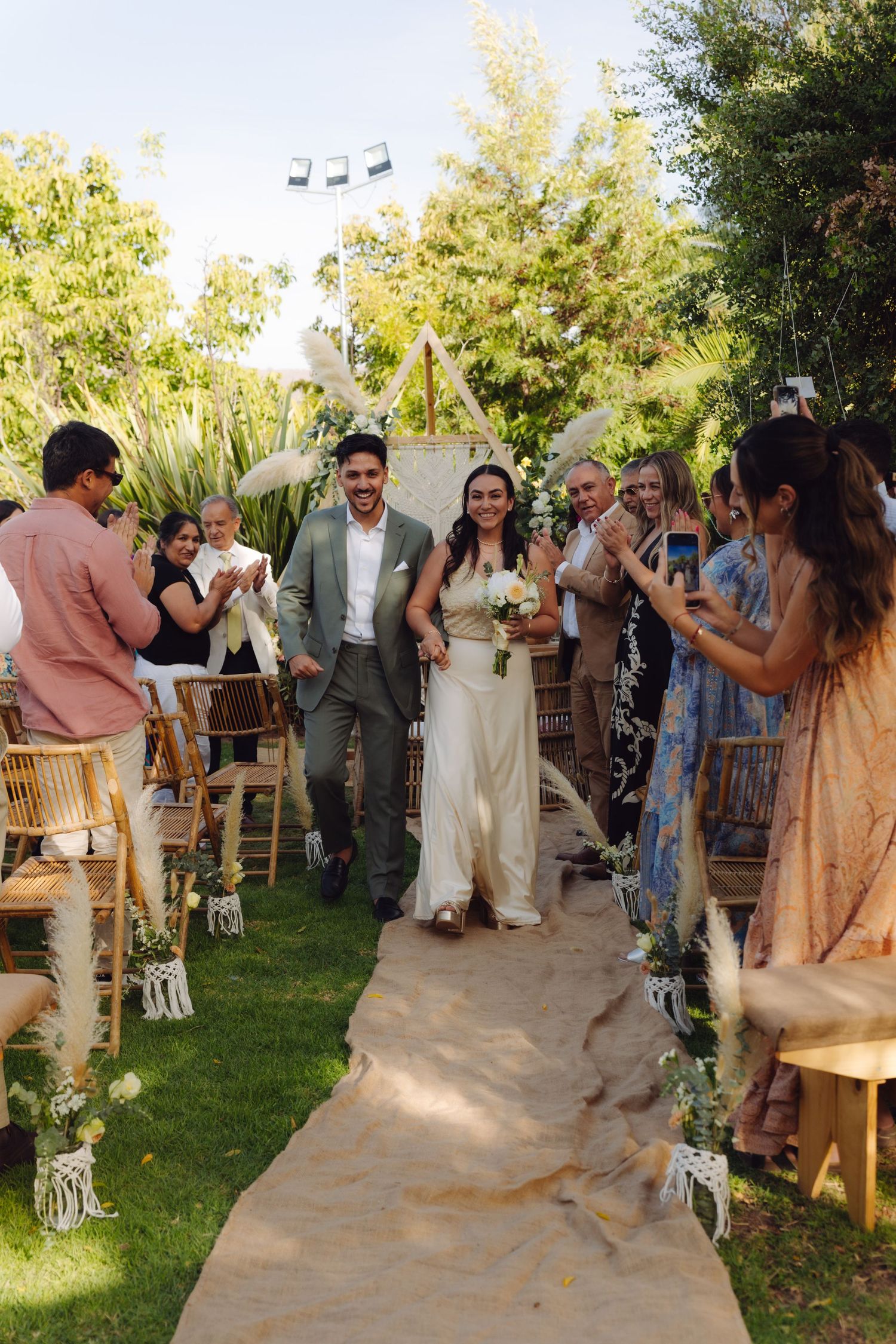 Smiling newlyweds walking down burlap aisle surrounded by cheering guests at outdoor wedding.