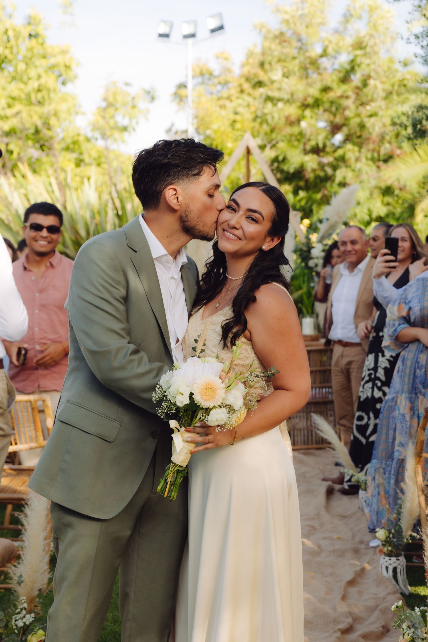 Groom kissing bride on cheek as she smiles holding white bouquet after outdoor wedding.
