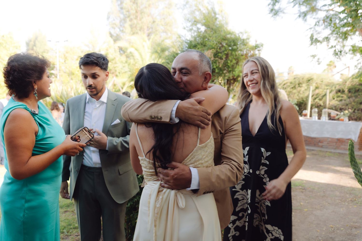 Wedding guests embrace warmly outdoors while smiling couple looks on at a sunny garden celebration.