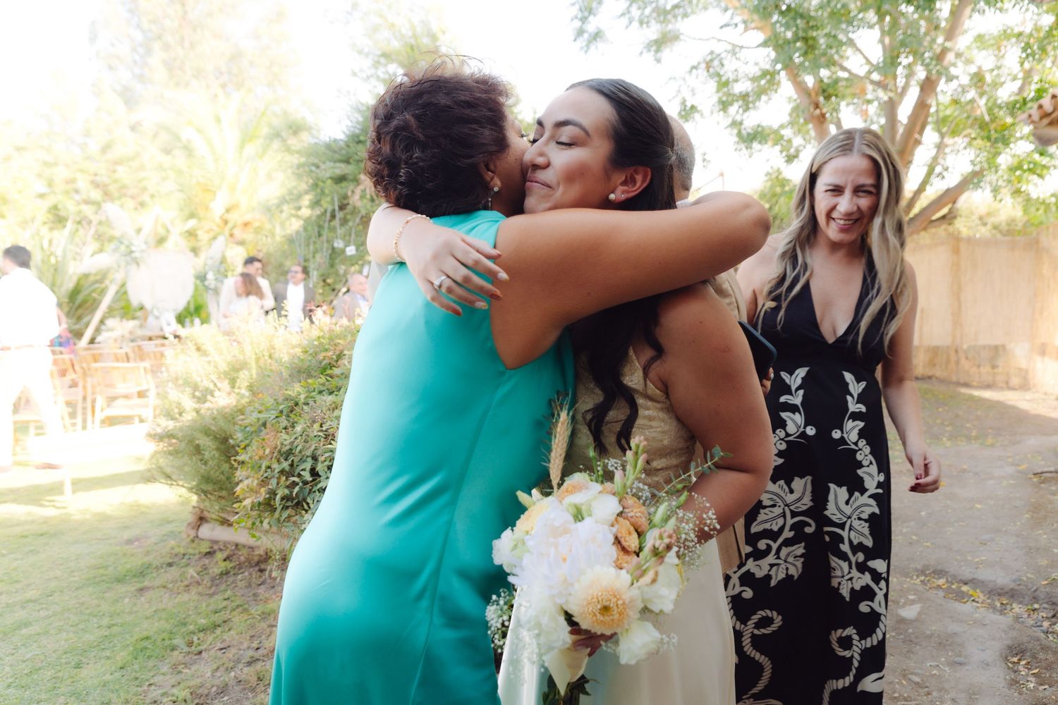 Bride holding bouquet receives warm hug from guest in teal dress at outdoor wedding reception.