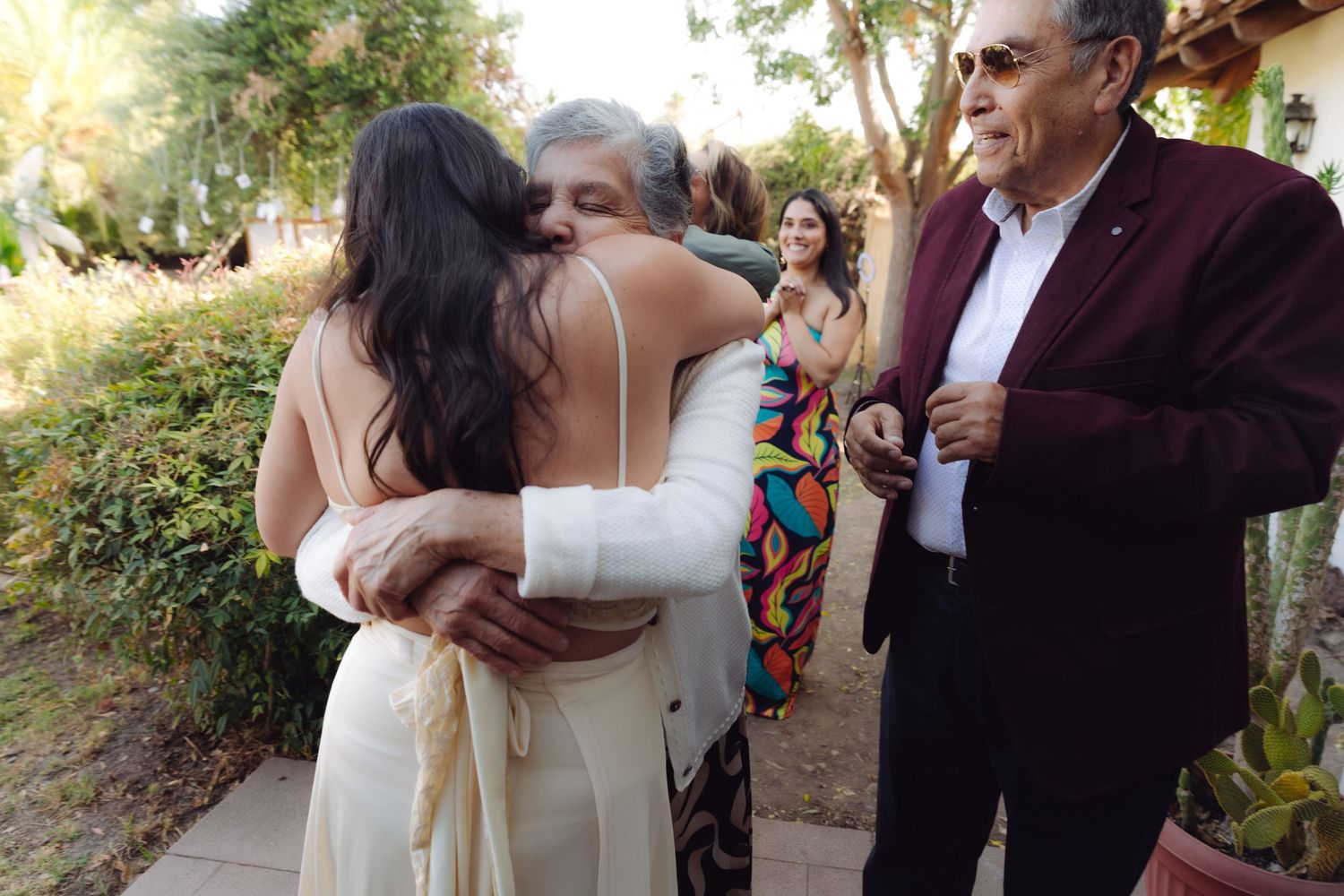 Bride in white gown shares heartfelt embrace with older guest at sunny outdoor wedding venue.