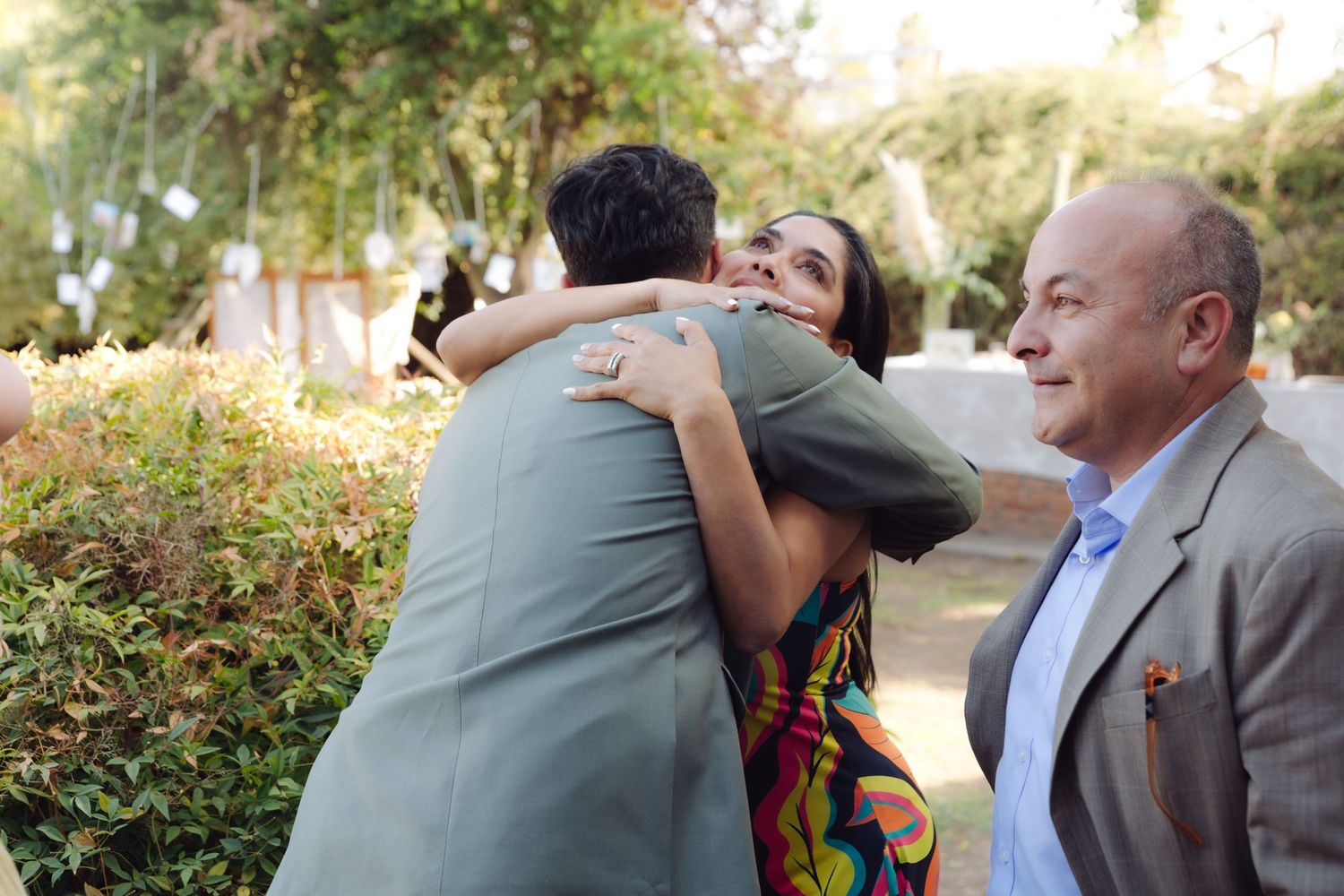 Wedding guest in colorful dress hugs groom in green suit while man smiles nearby outdoors.