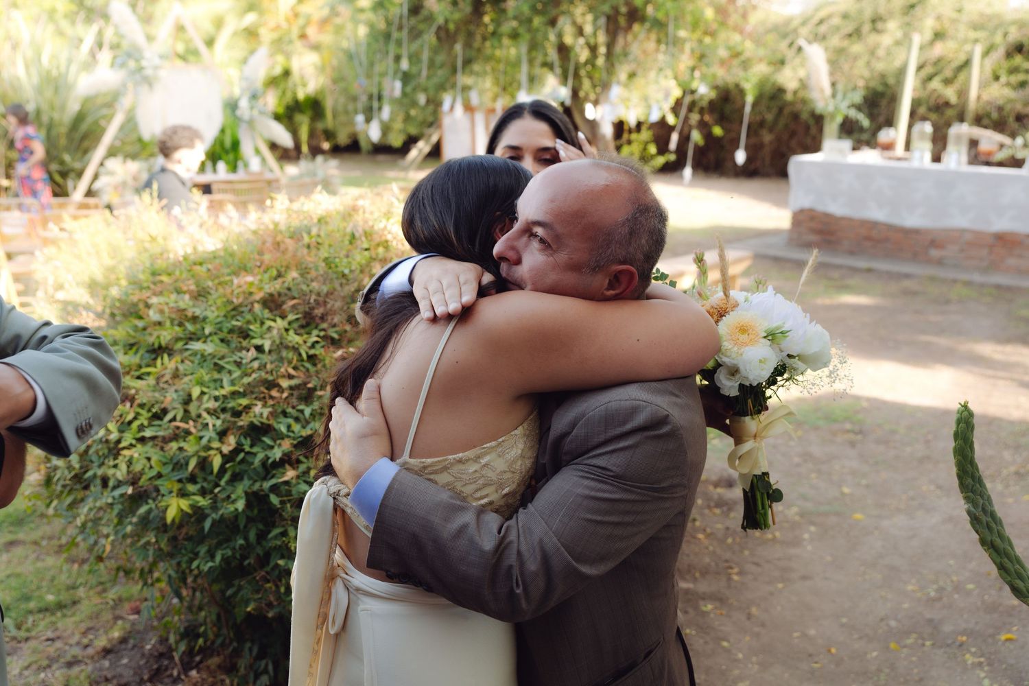 Emotional wedding embrace between bride holding white bouquet and suited guest in garden setting.