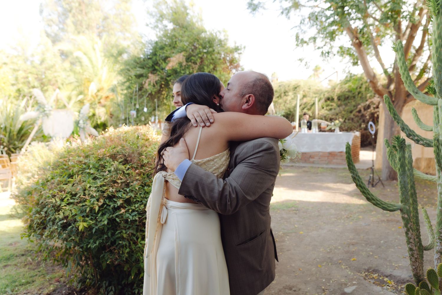 Bride and groom share tender three-way hug with wedding guest among lush garden greenery.