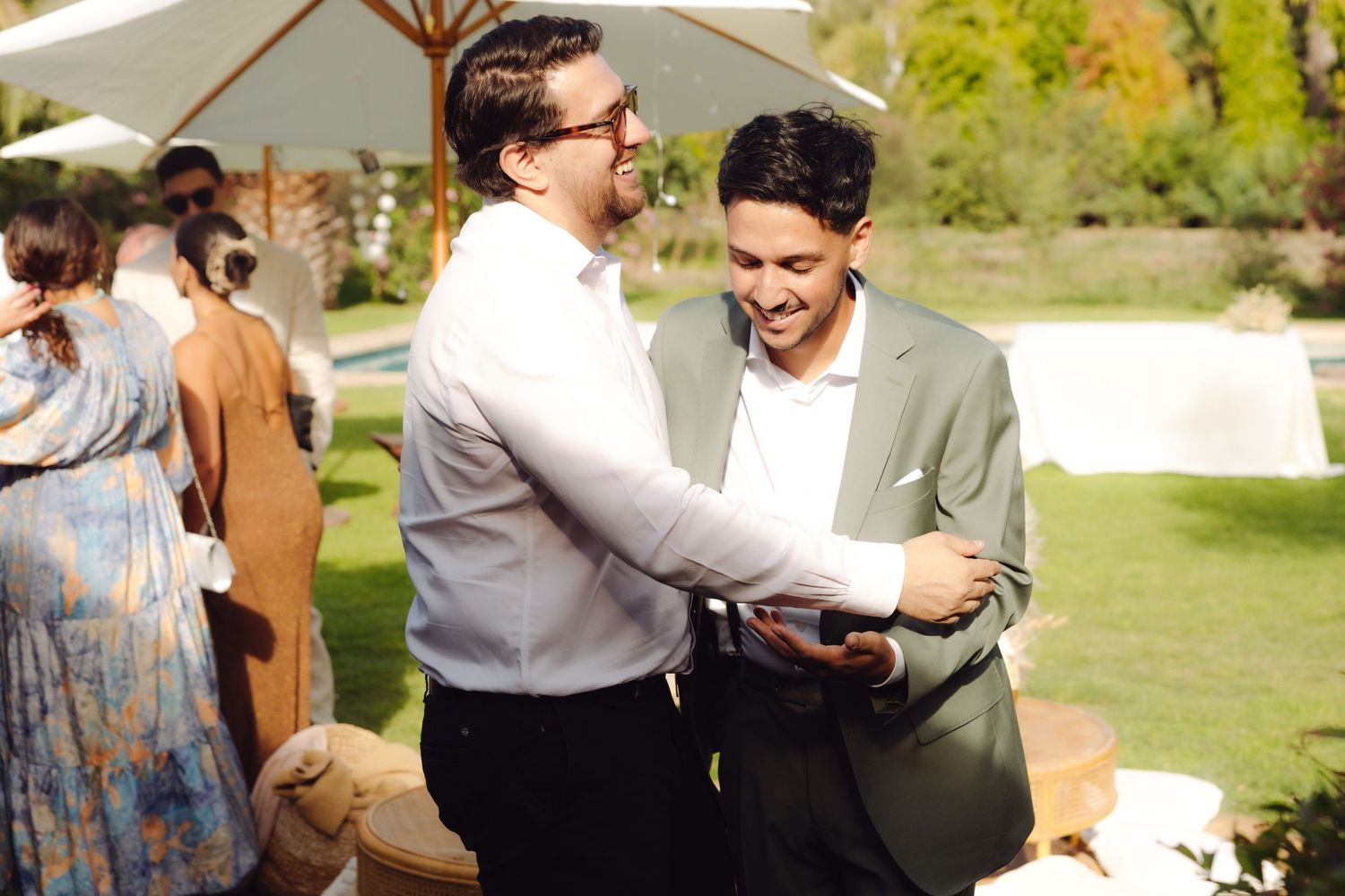 Two men laugh and share a friendly handshake at an elegant outdoor wedding reception.