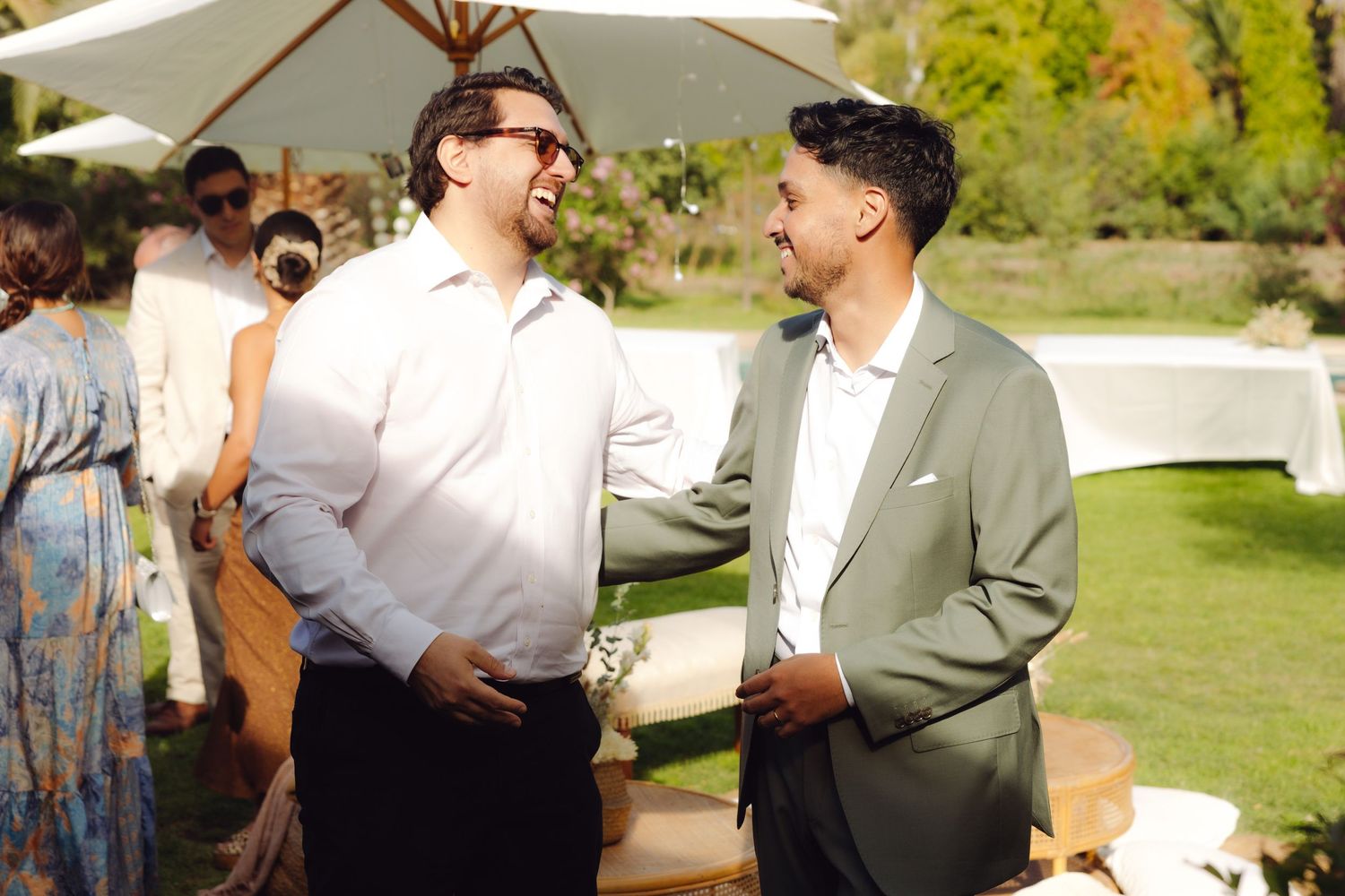 Two well-dressed men smile and chat together at a sunny outdoor wedding celebration.