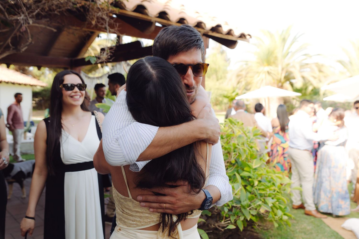 Bride receives warm hug from sunglasses-wearing guest at lively outdoor wedding reception.
