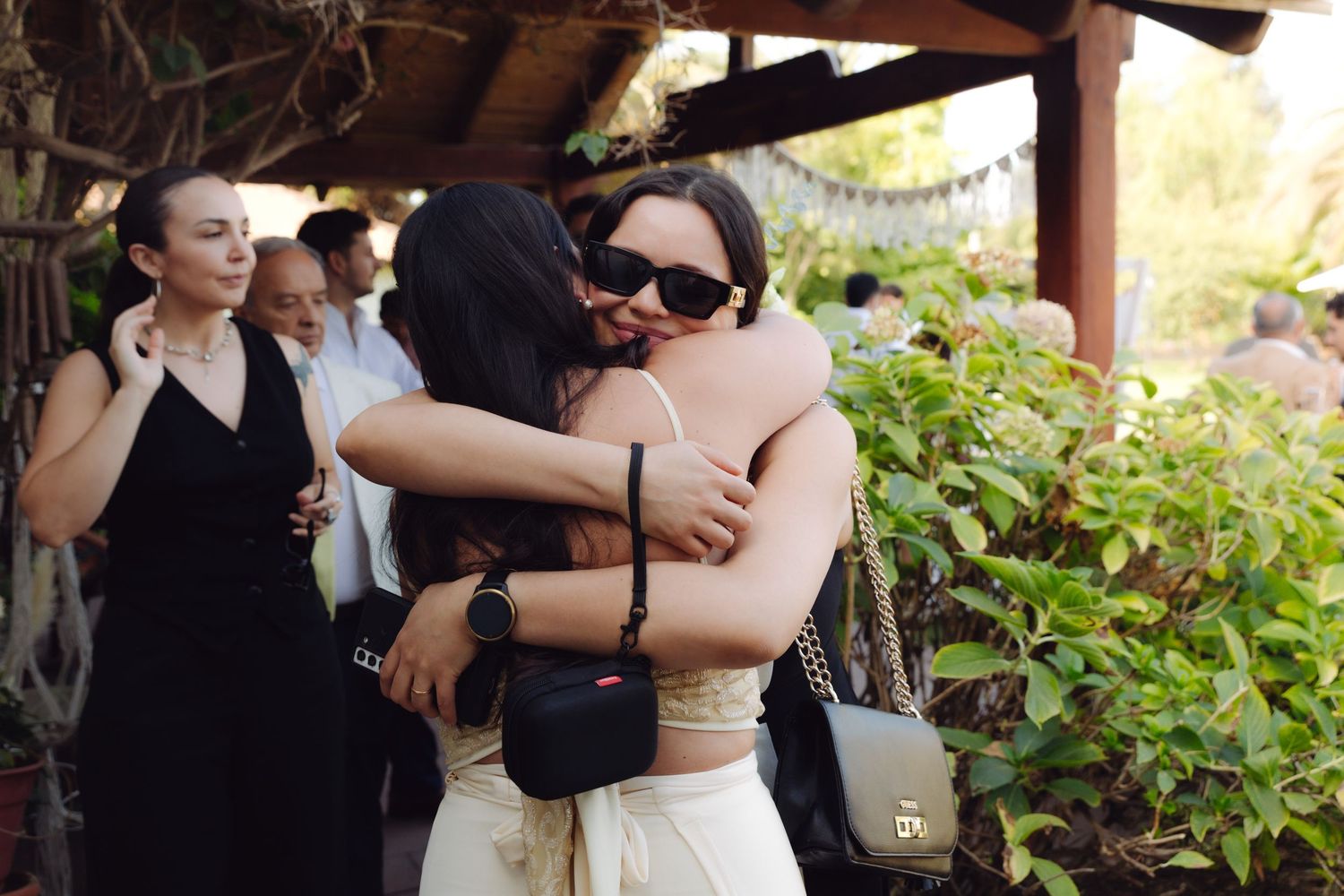 Two women share an emotional embrace at an outdoor wedding while guests mingle nearby.